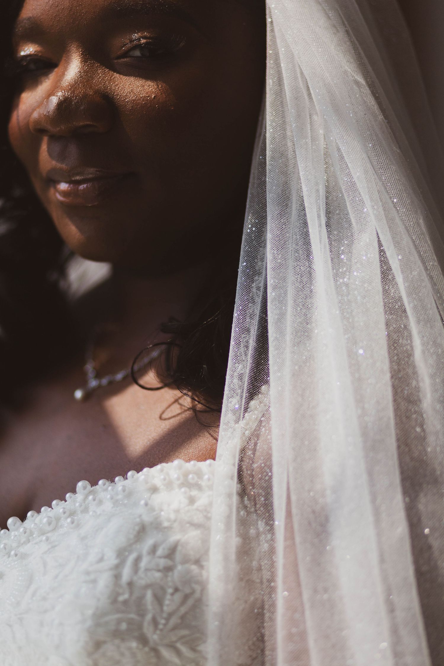 Close-up of a bride in a beaded white lace wedding gown and sparkling tulle veil, softly lit by warm sunlight.