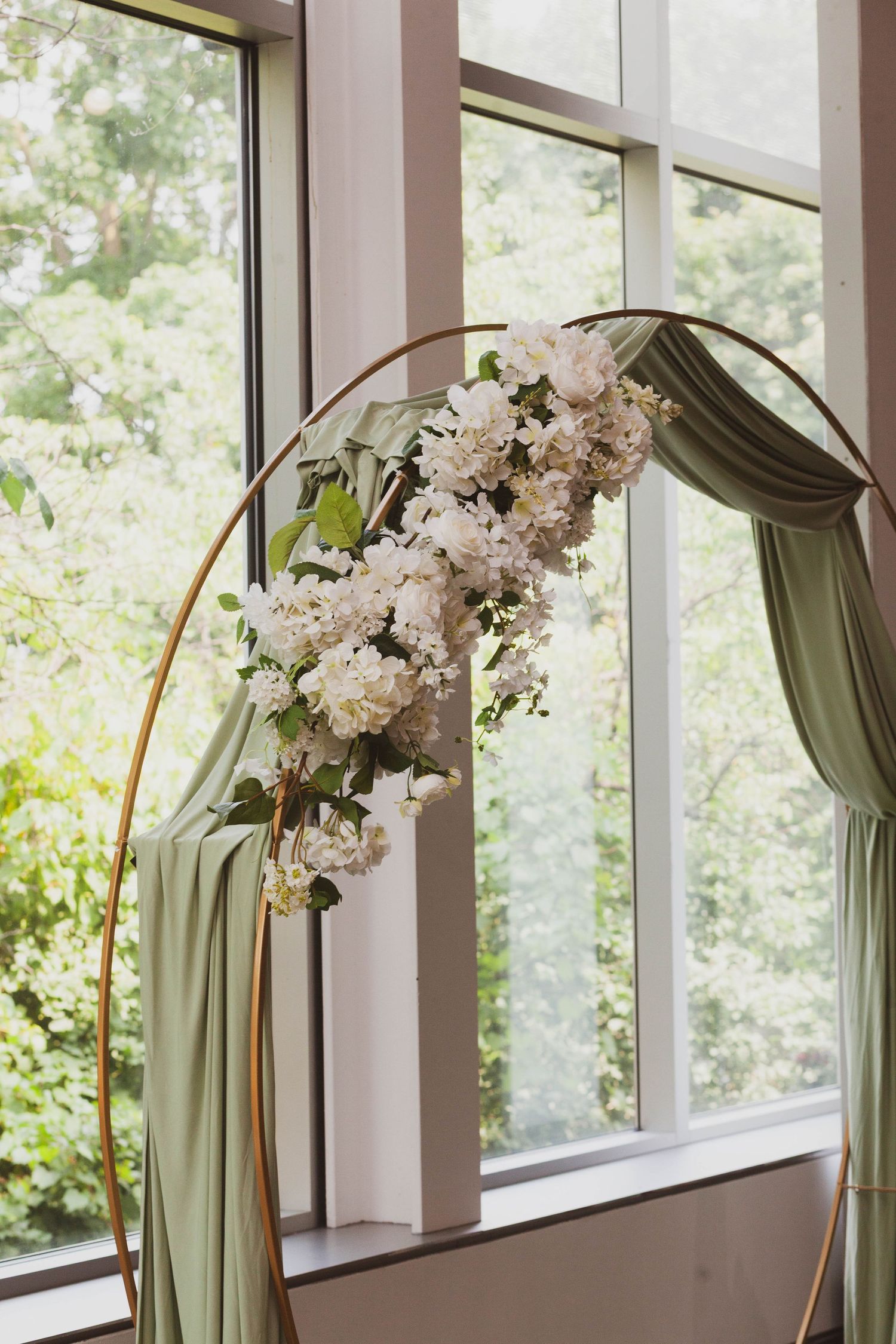 Round gold wedding arch draped with sage green fabric and white floral arrangement, set before large windows overlooking lush greenery.