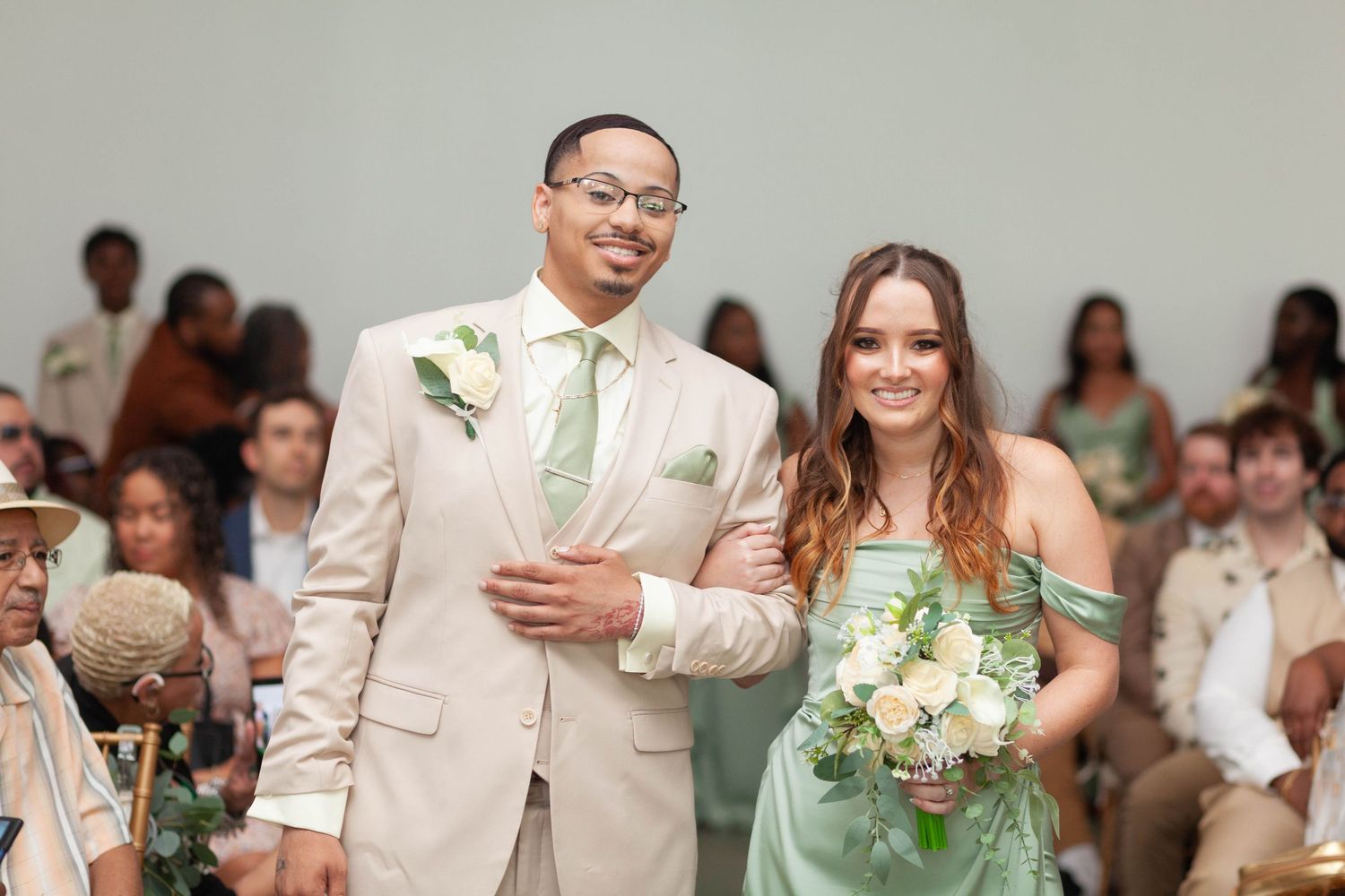 Smiling groomsman in a tan suit with a sage tie walks arm in arm with a bridesmaid in a green dress holding white flowers as guests watch.