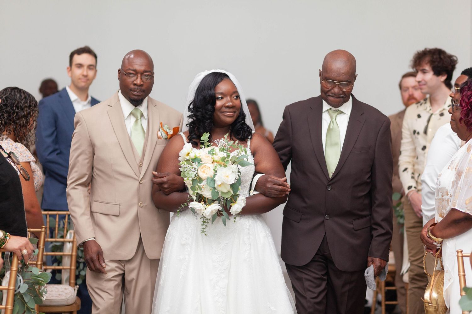 A bride in a white gown holding a bouquet of white flowers walks down the aisle arm in arm with two men in suits as guests look on.