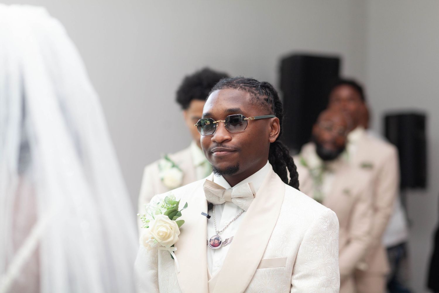 Groom in a cream tuxedo, bow tie and tinted glasses stands at the altar with a white floral boutonniere as groomsmen look on behind him.