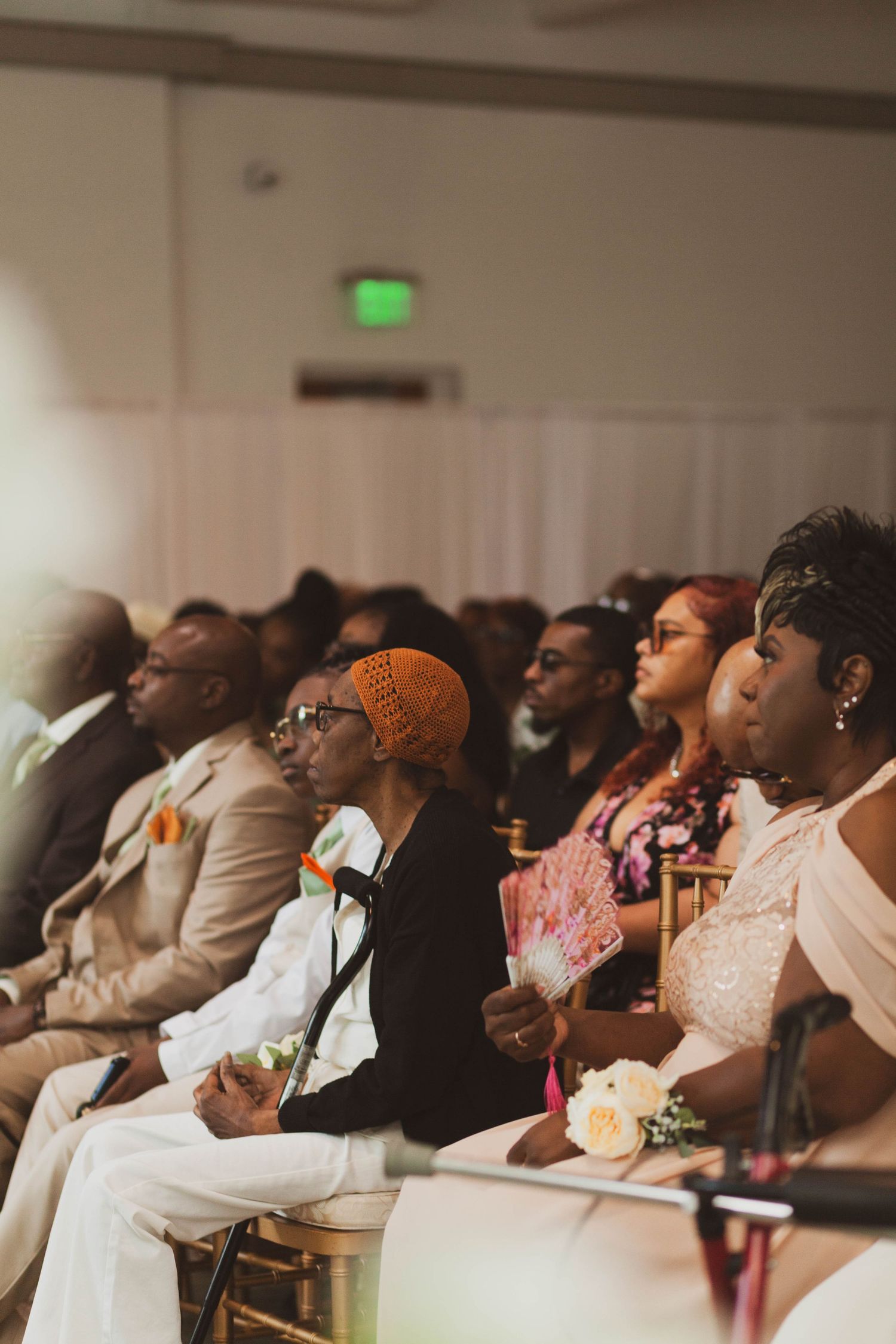 Wedding guests in formal attire sit on gold chairs, watching a ceremony in a bright banquet hall, with one woman holding a floral fan.