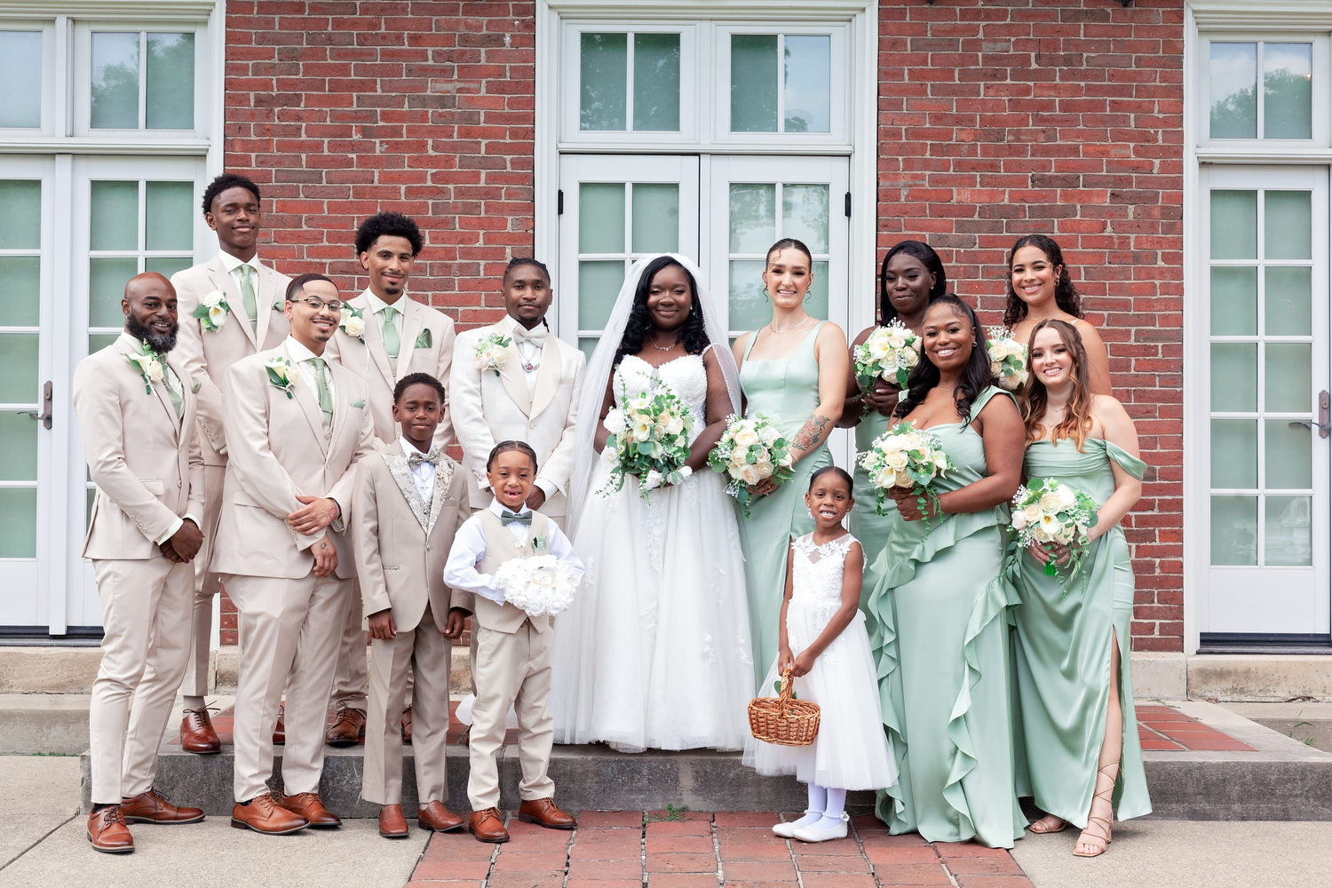 Bridal party in sage green and tan poses with the bride and flower girl on brick steps outside a white-trimmed building.