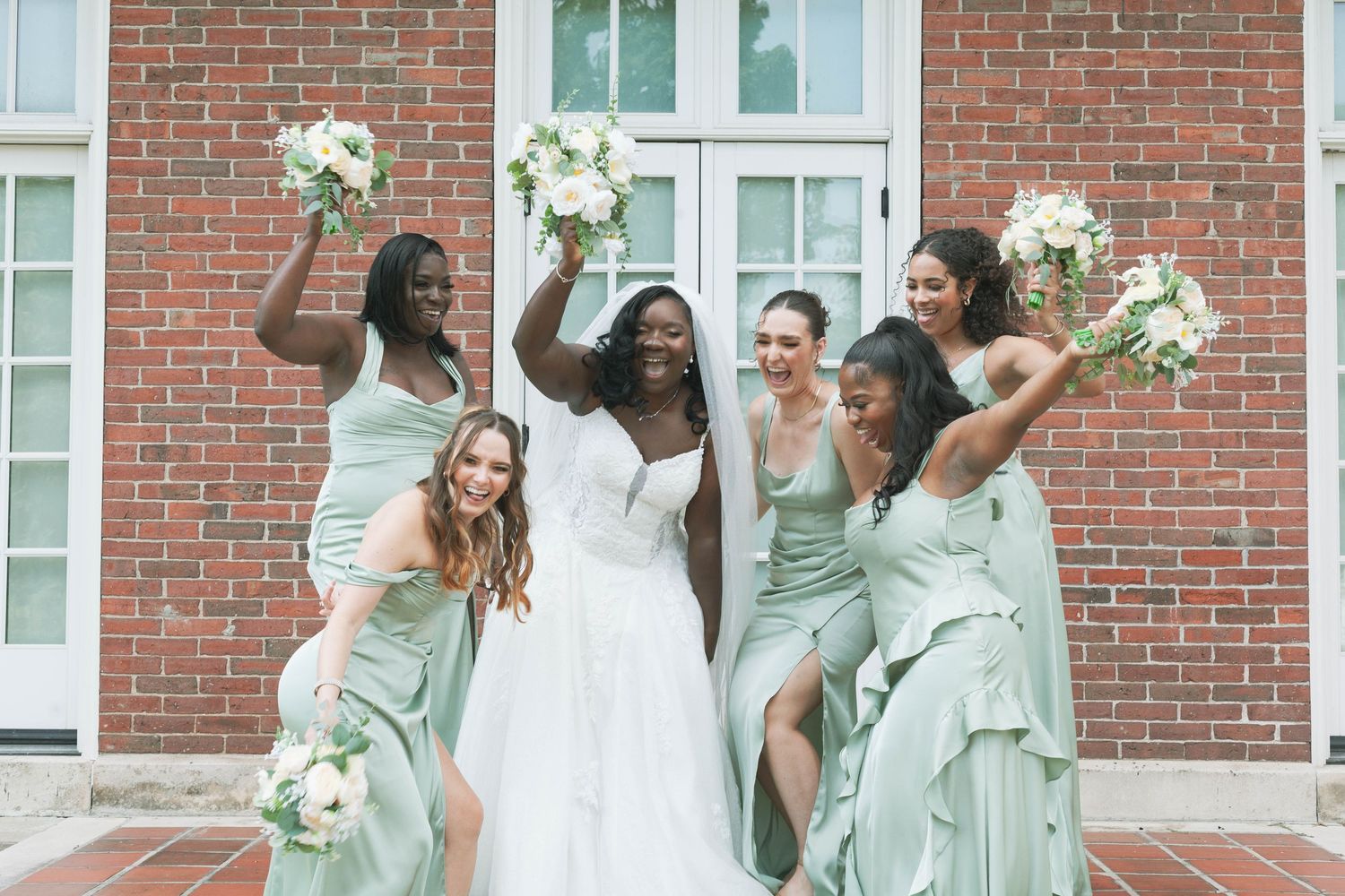 Smiling bride in a white gown celebrates with five bridesmaids in sage green dresses holding bouquets in front of a red brick building.