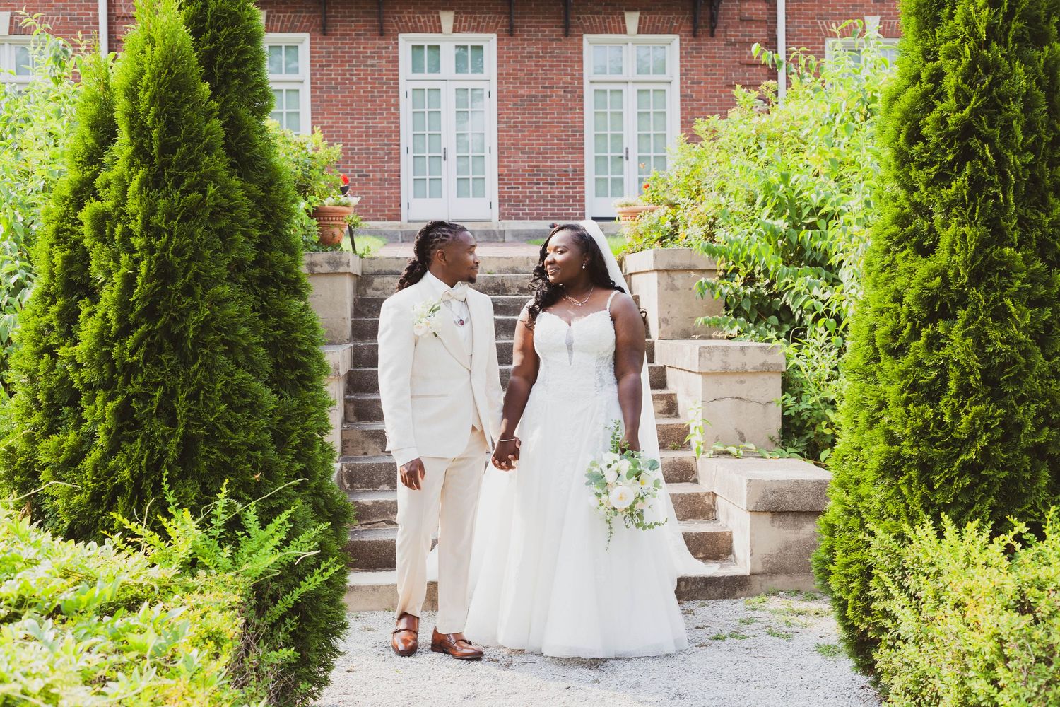 Bride in a white gown holding a bouquet and groom in a cream suit hold hands on garden steps beside evergreen shrubs and a brick building.