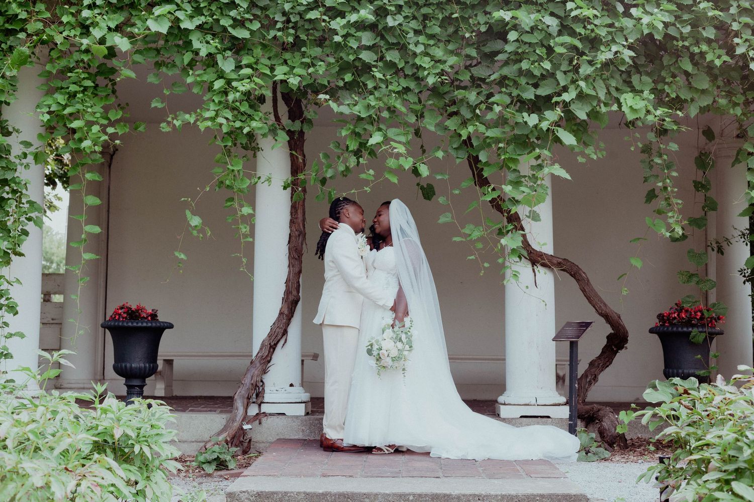 A bride in a long white gown and veil embraces her groom in a light suit beneath vine-covered columns at an outdoor wedding venue.