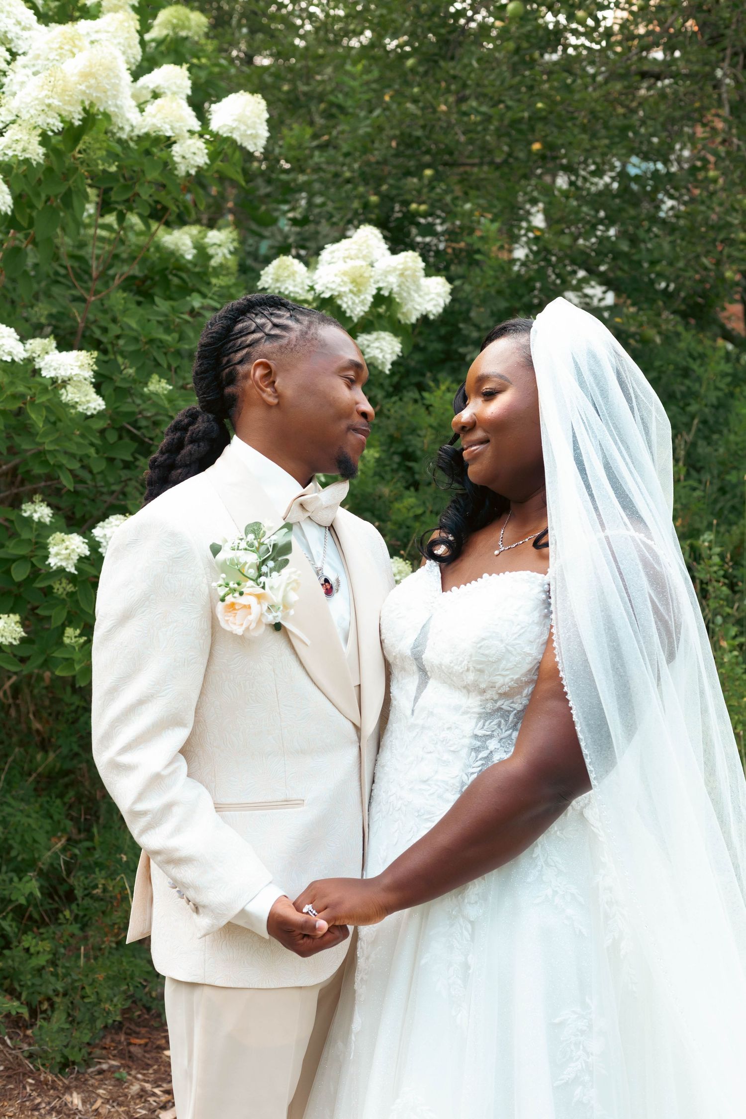 A smiling bride in a white lace gown and veil holds hands with a groom in a cream suit and floral boutonniere beside blooming white flowers.