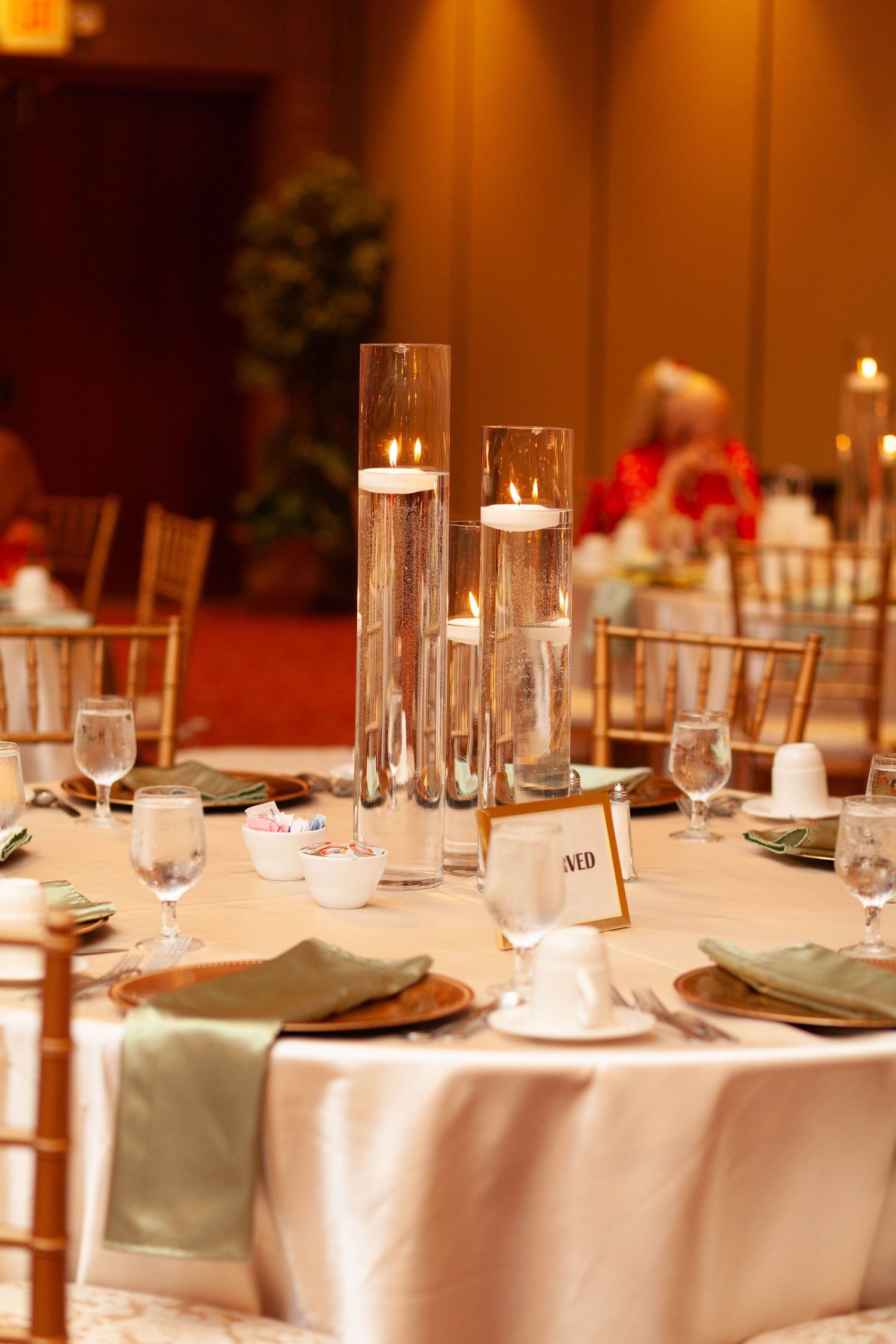 Elegant banquet table with floating candles in tall glass cylinders, green napkins, wine glasses, and place settings in a ballroom.