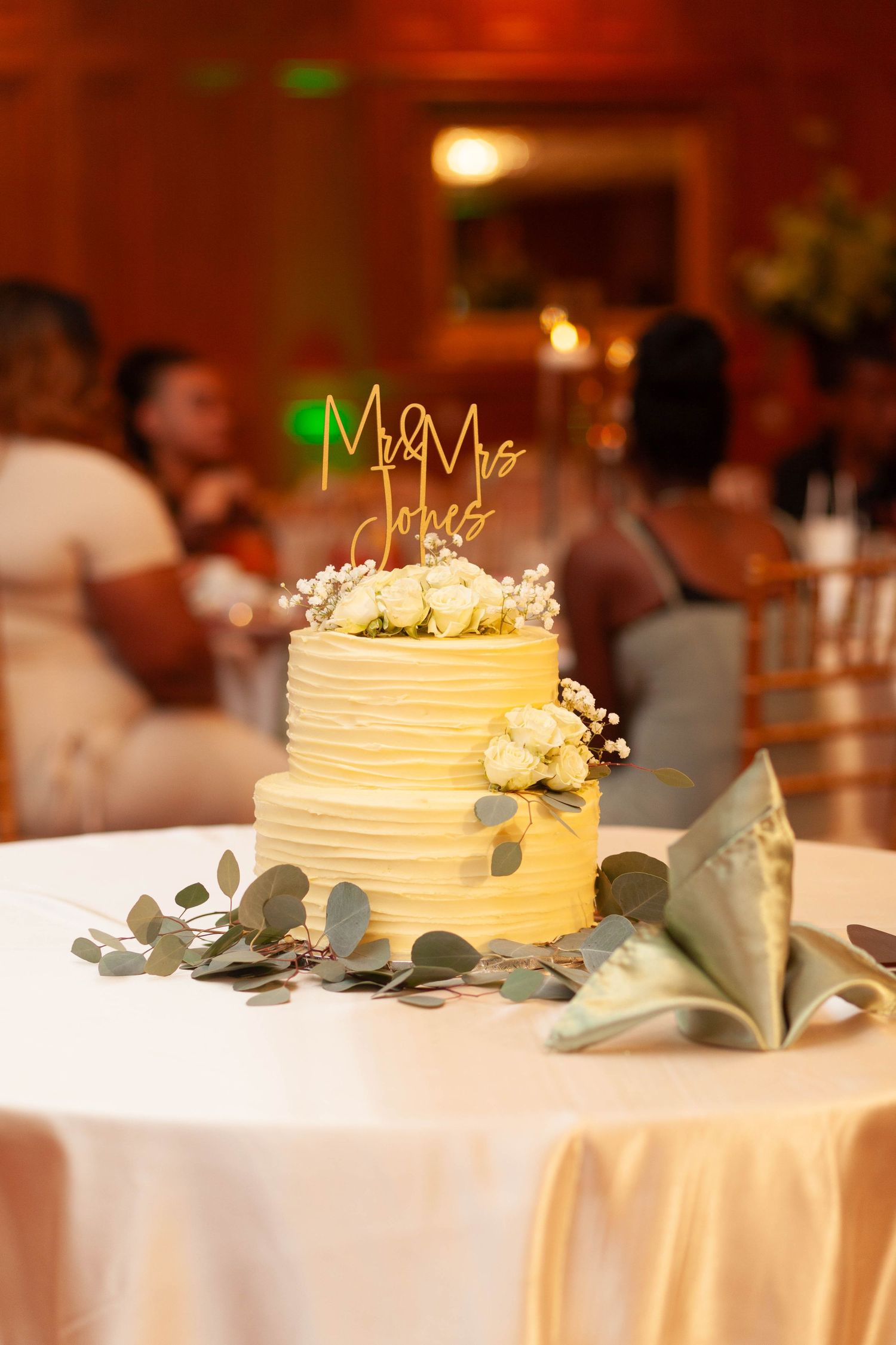 Two-tier white wedding cake with a gold Mr and Mrs Jones topper and white flowers on a table with eucalyptus and a green napkin.