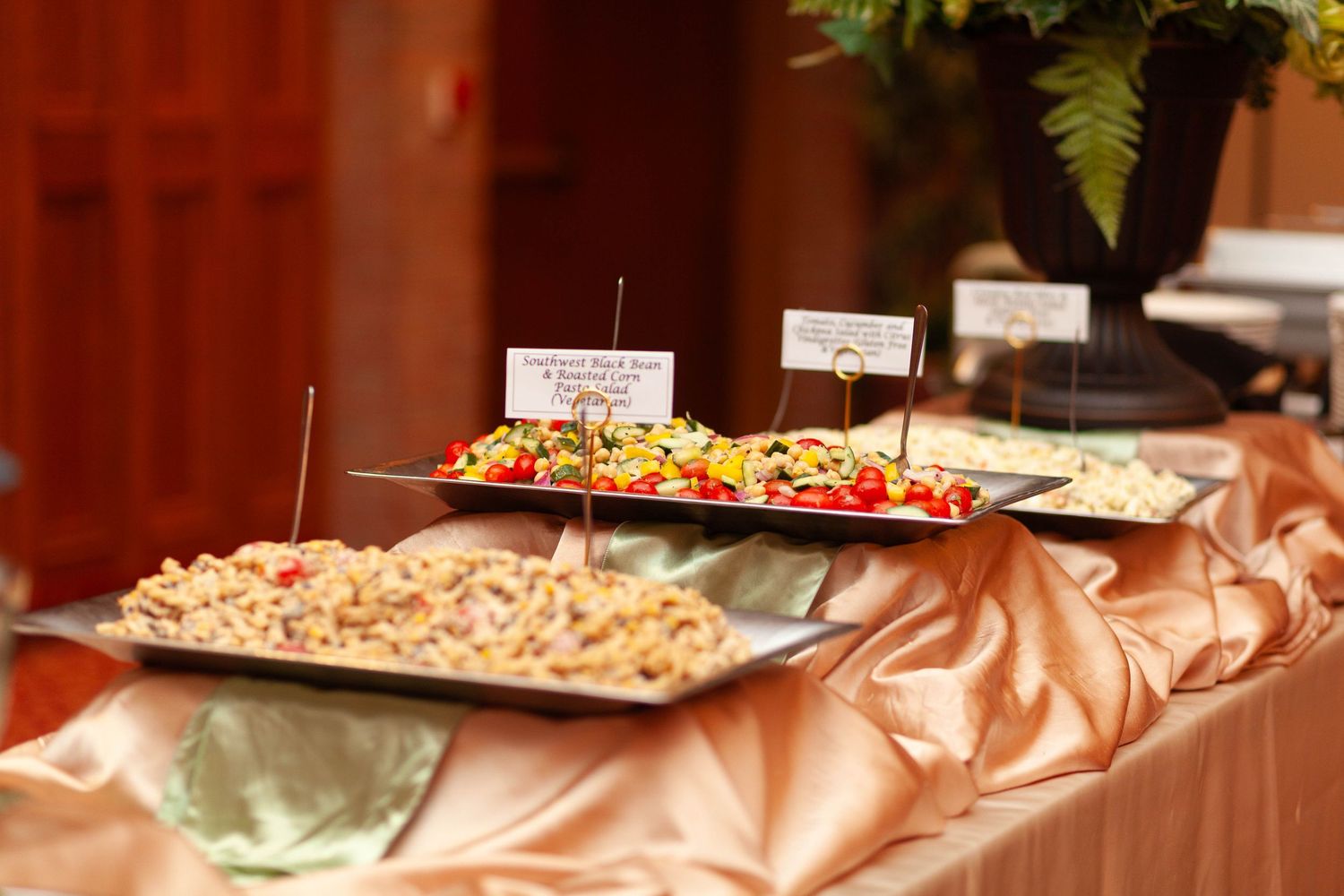 Buffet table draped in satin with silver trays of colorful pasta salads, labeled with small signs, in an event hall.