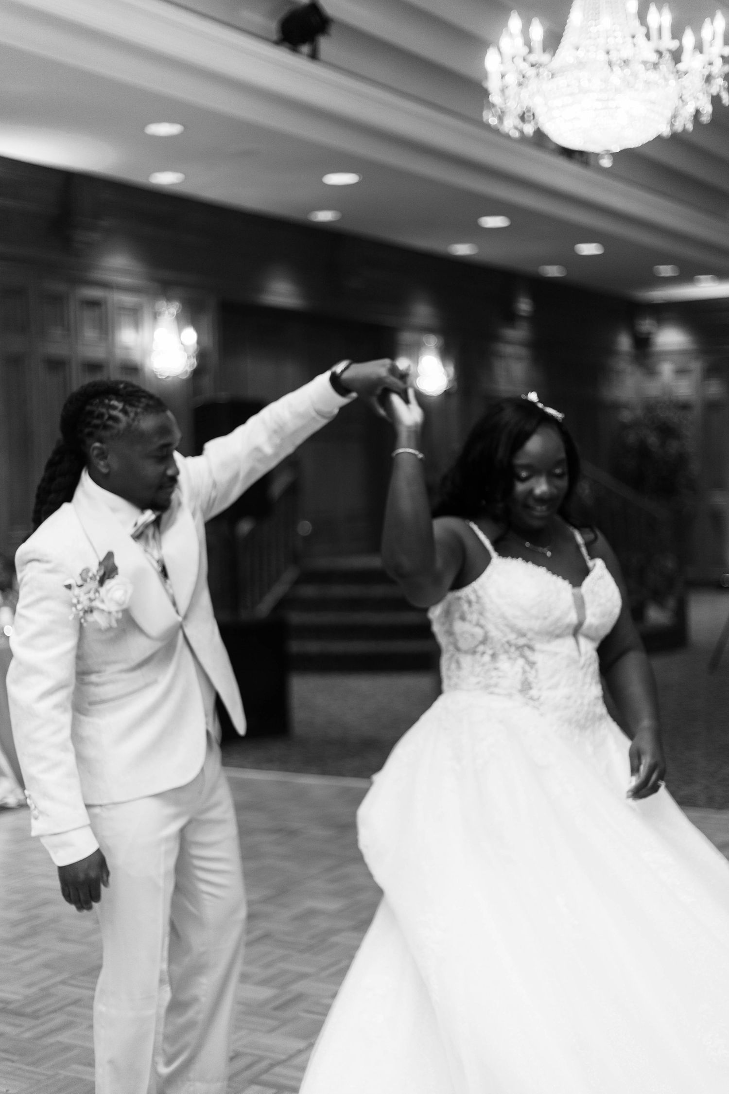 Black and white photo of a bride in a lace ball gown twirling under her partner's raised arm during a first dance in an elegant ballroom.