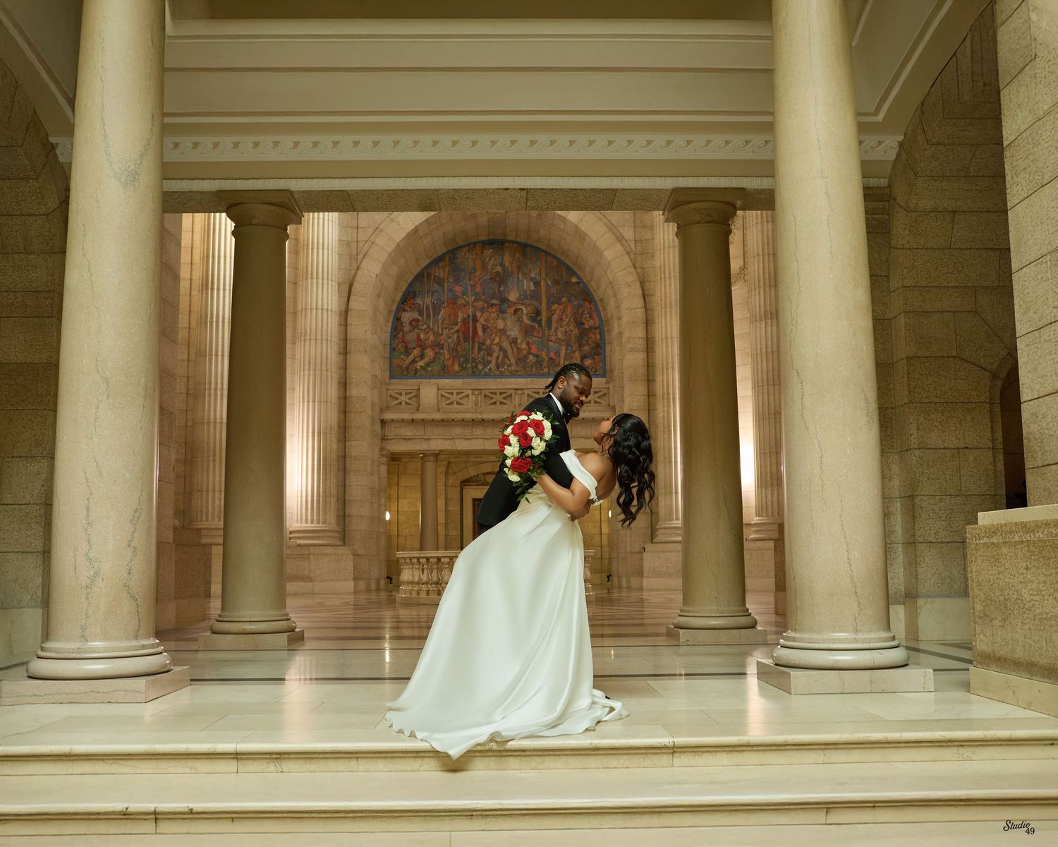 Smiling bride in a white gown holding red and white roses sits beside a groom in a black tuxedo and sunglasses in a vintage convertible.