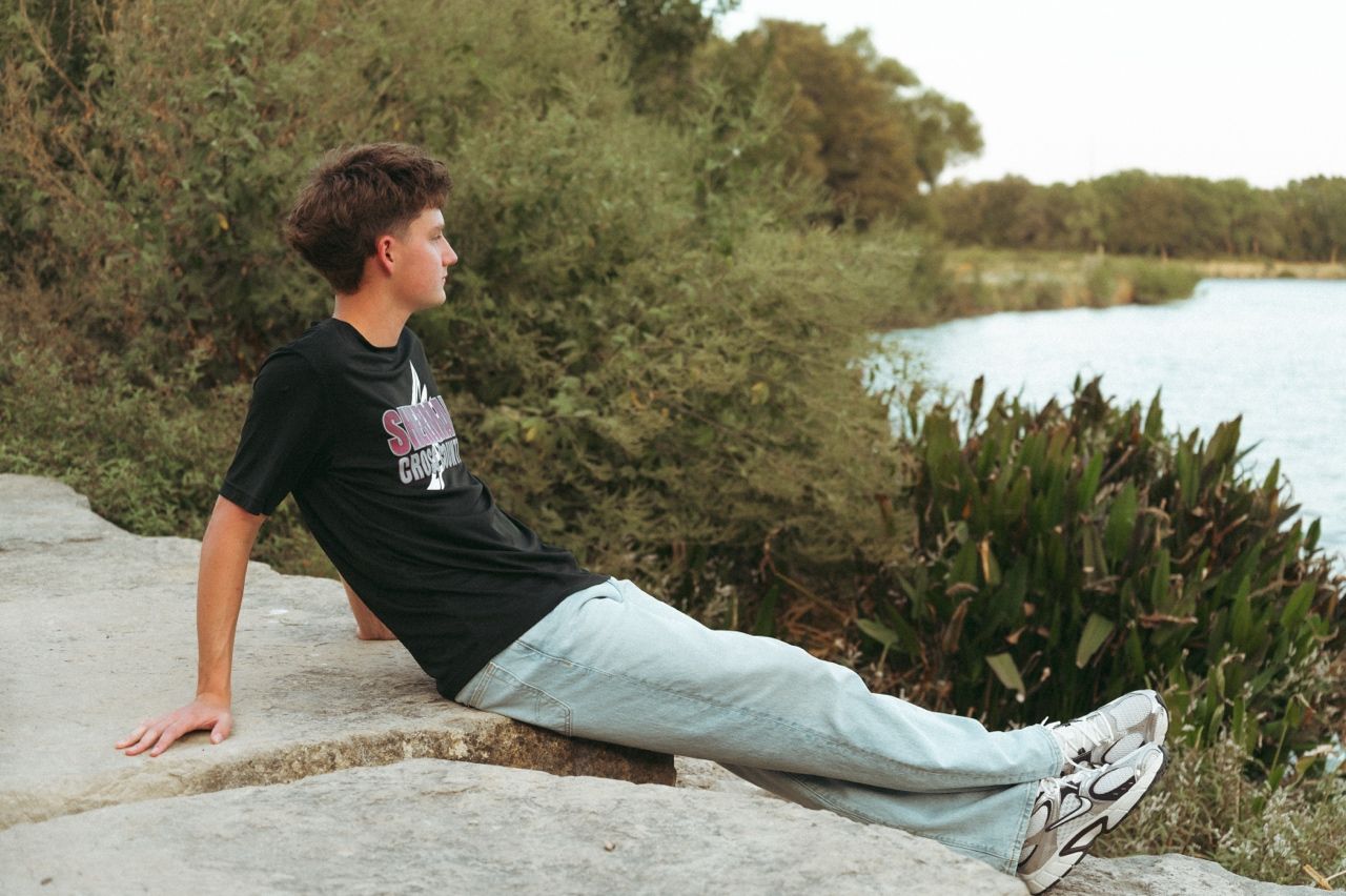 Teen boy in black graphic tee and light jeans sits on rocks by a pond at Oak Point Nature Preserve.