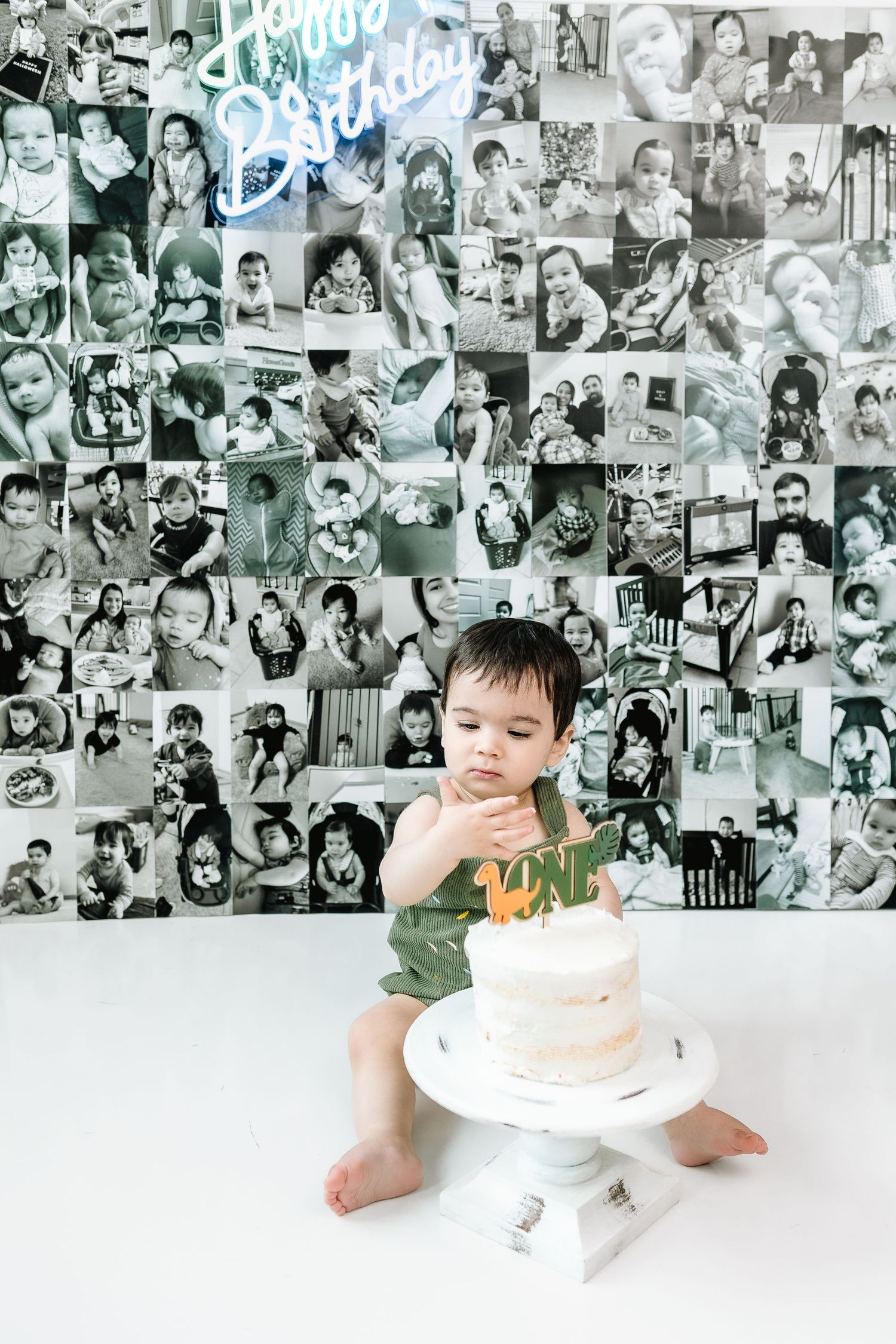 Baby boy in green outfit touches first birthday cake, surrounded by black and white photo collage wall with Happy Birthday sign.