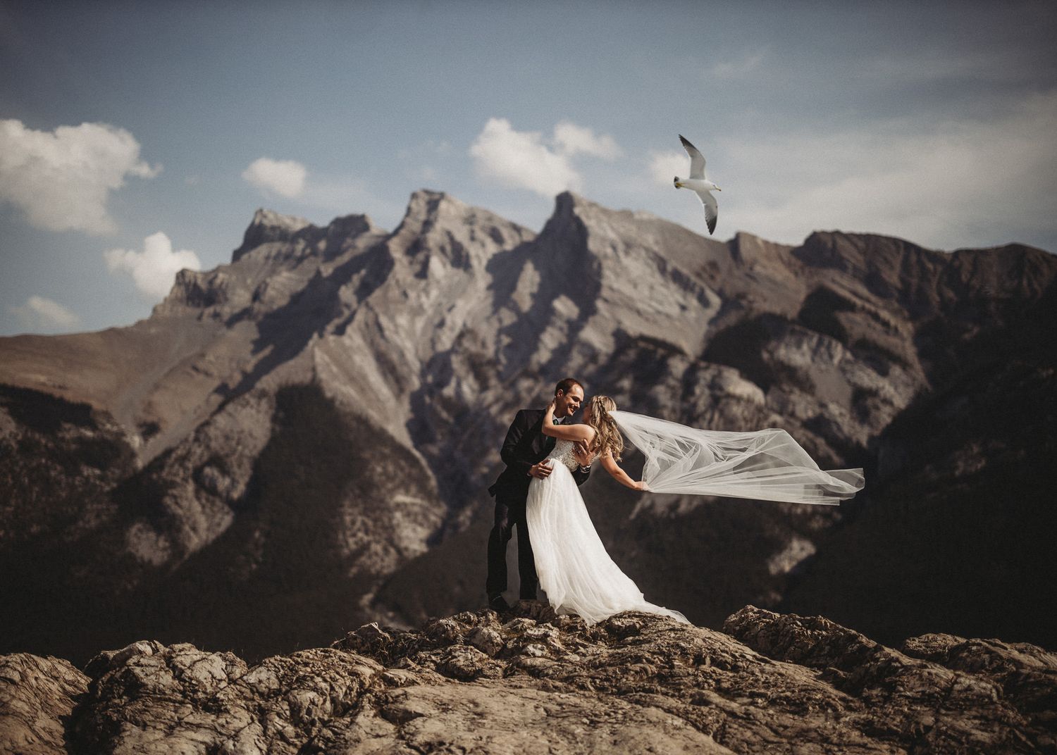 Bride and groom kissing on a rocky mountain summit as her veil streams in the wind, by a Banff wedding photographer