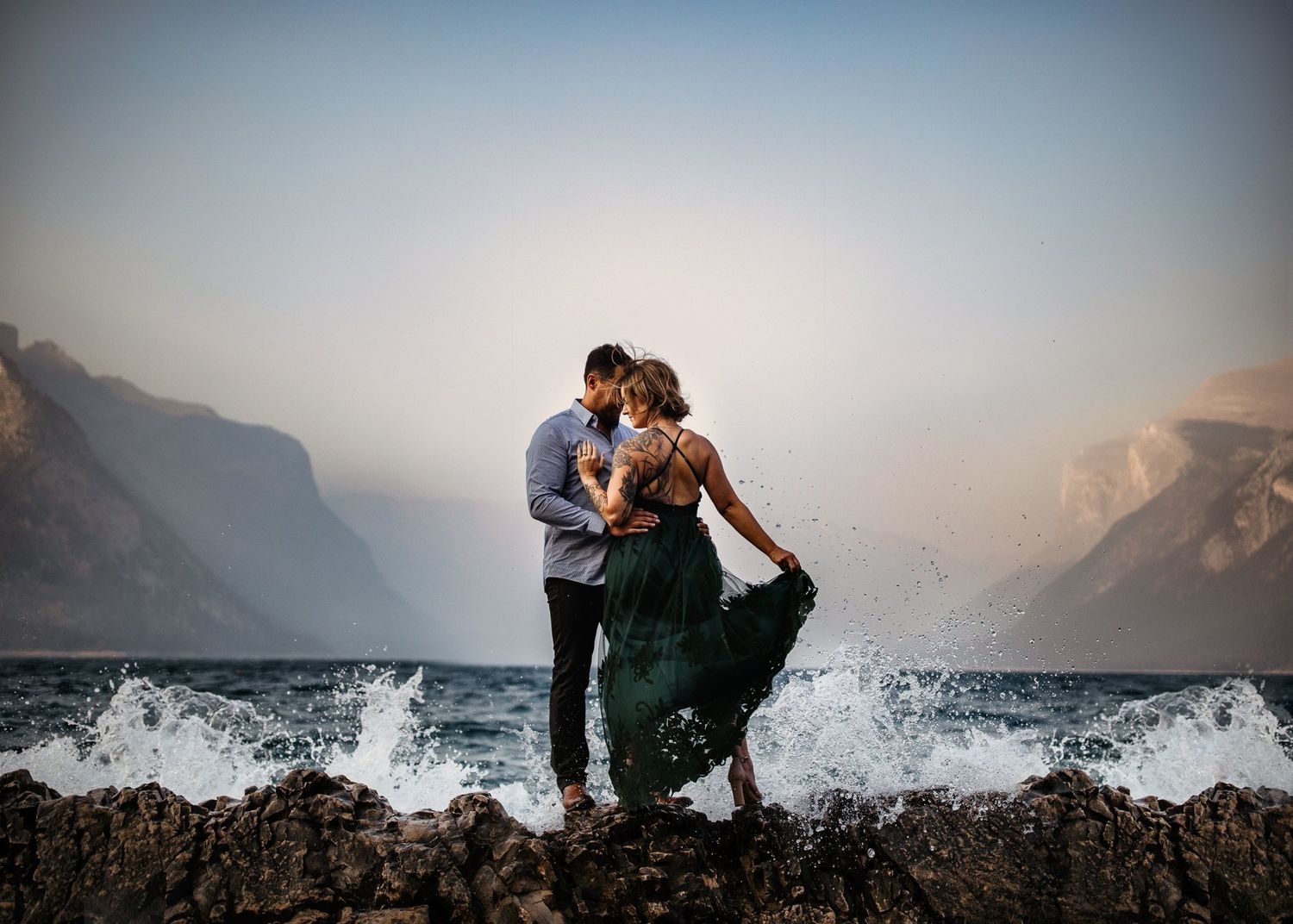 Bride in an emerald gown and groom on a misty mountain lakeshore as waves crash, by a Lake Louise wedding photographer