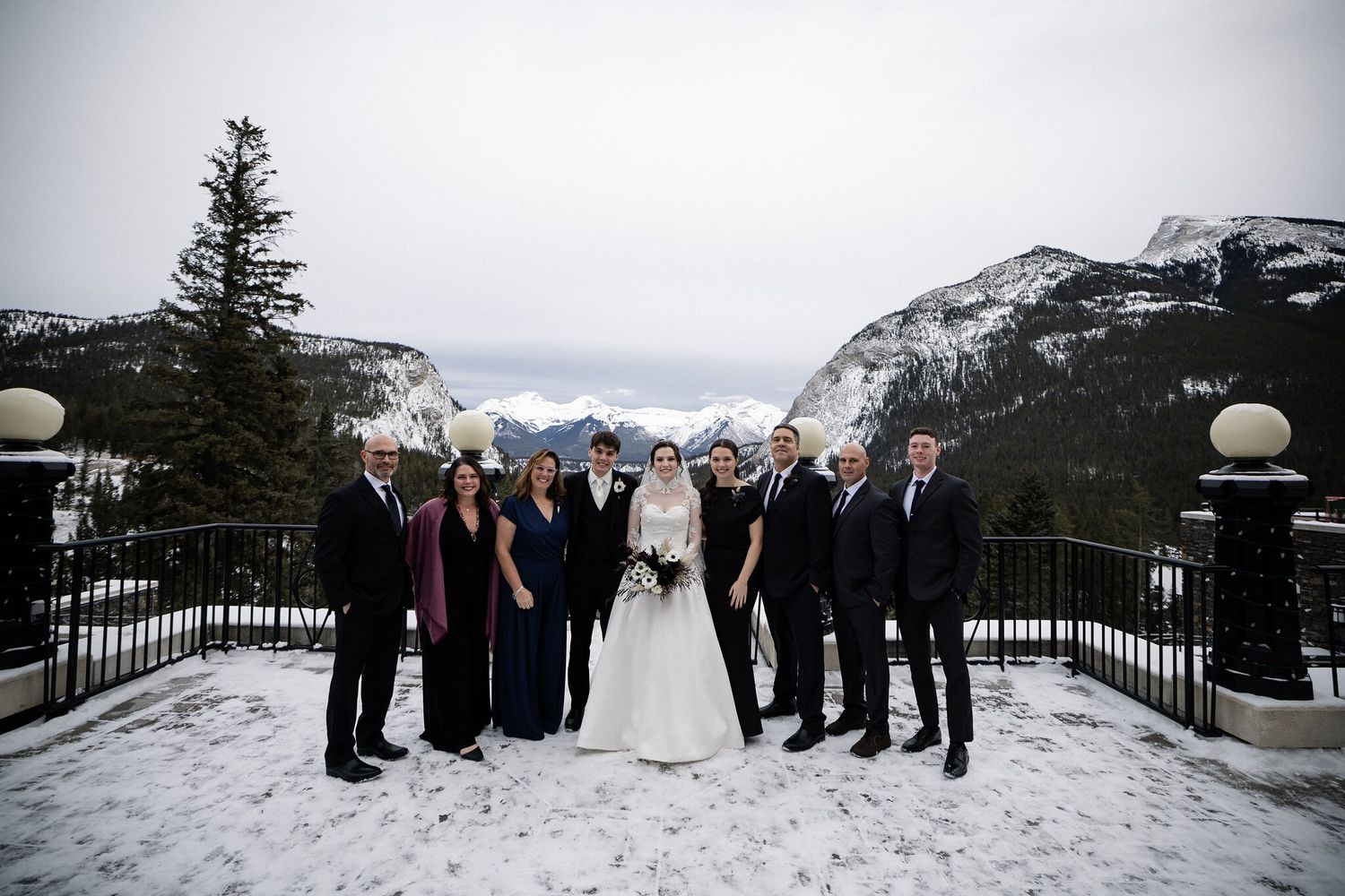 Wedding group photo on the snowy Fairmont Banff Springs terrace framed by mountains and globe lamp posts