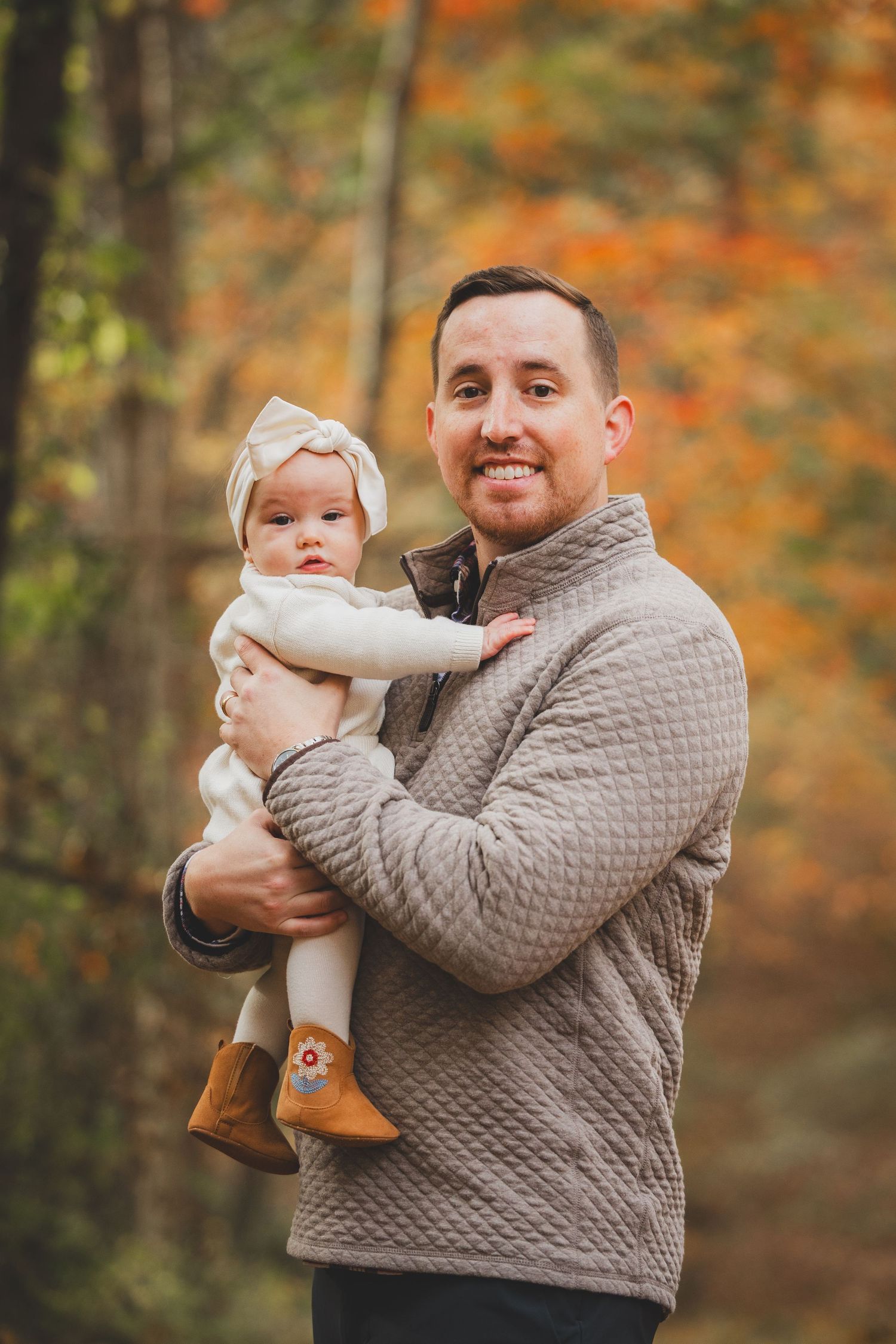 A smiling man holds a baby girl in a white outfit and bow headband outdoors during autumn at Lake Fairfax Park