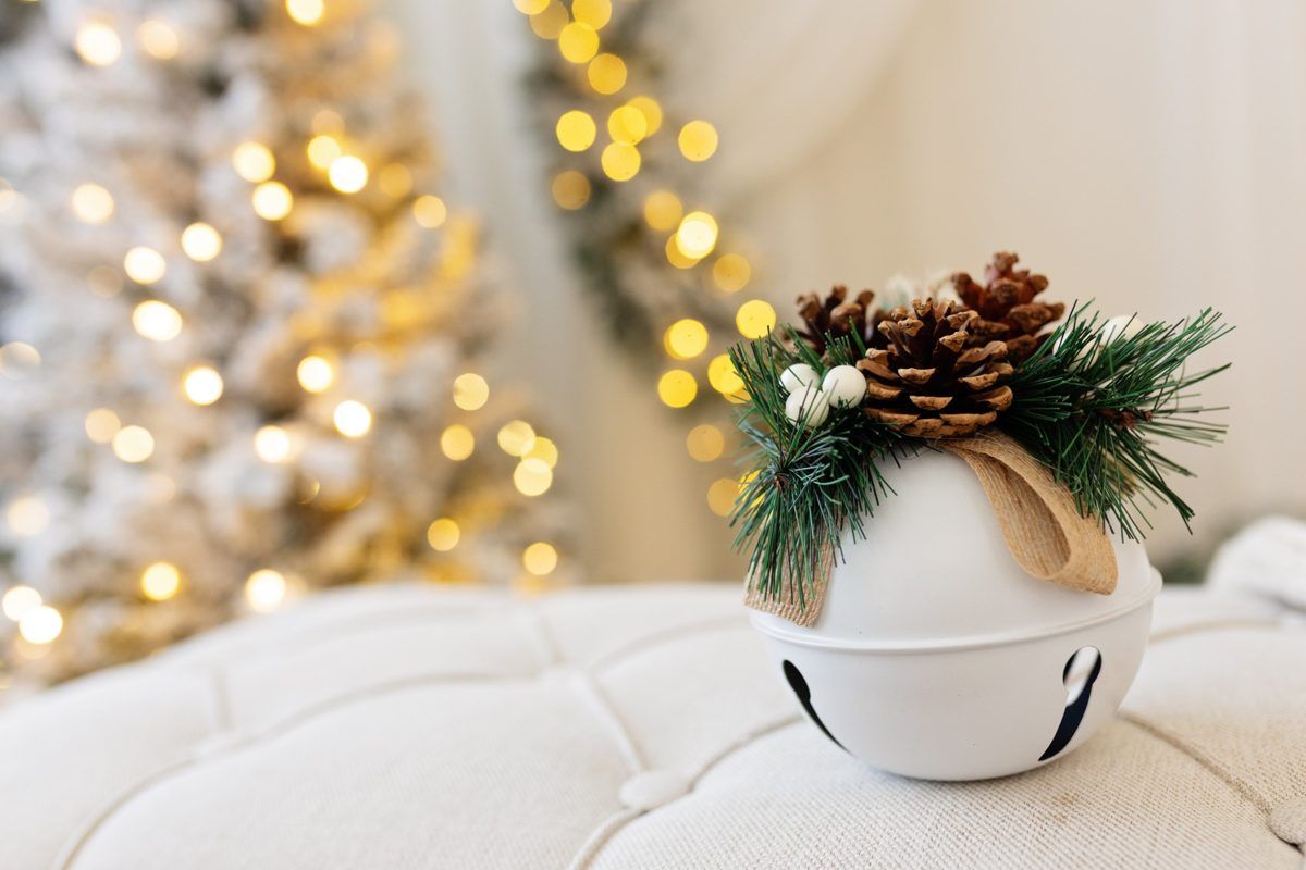 White jingle bell ornament decorated with pine cones and evergreen sprigs on a tufted seat, with a glowing Christmas tree in the background.