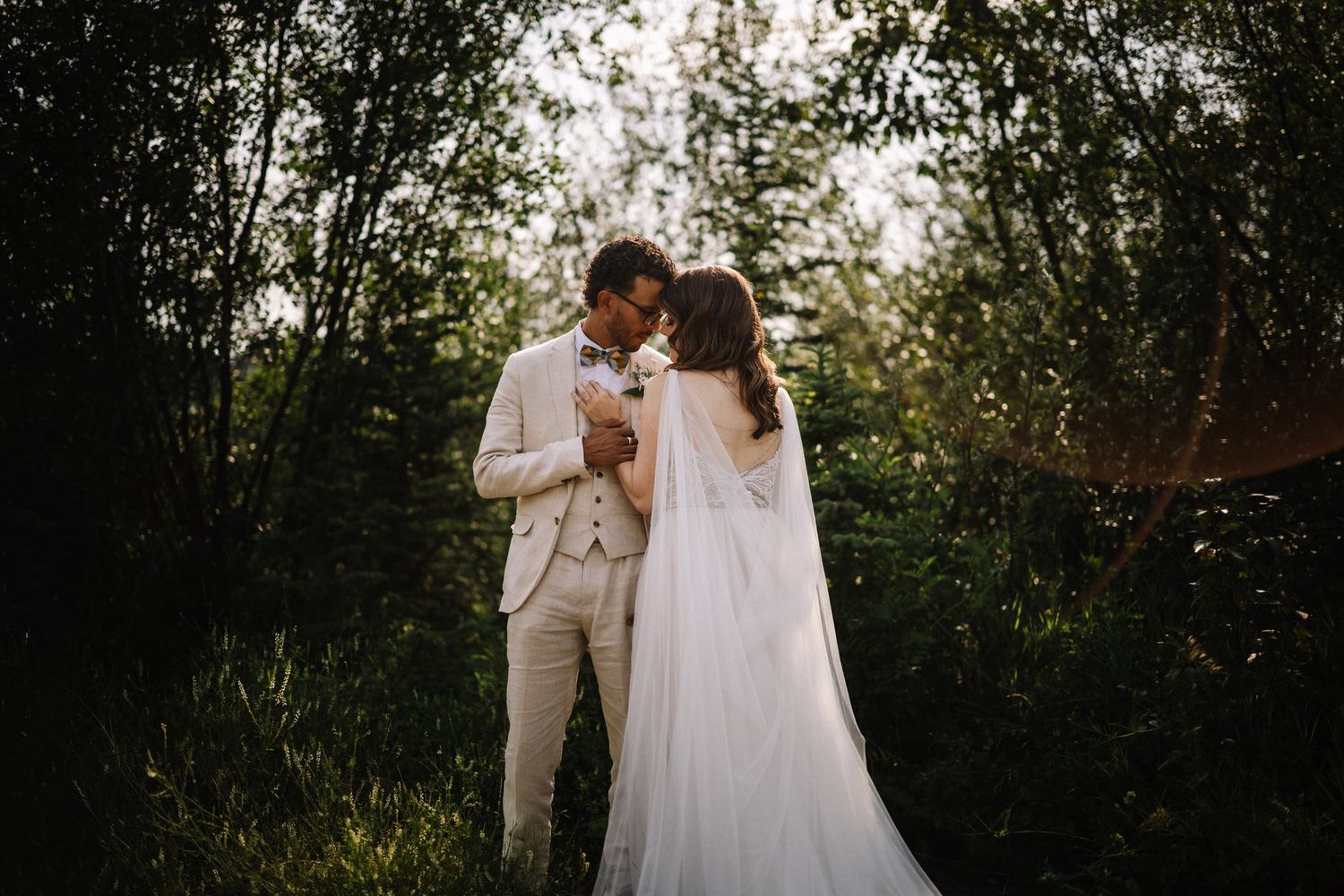 Couple standing close in deep green trees with the bride's long veil trailing