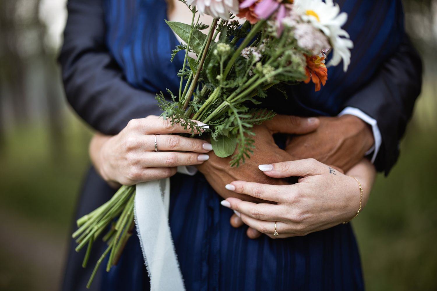 Close-up of a wildflower bouquet held against a navy dress, his arms wrapped around her and engagement ring showing