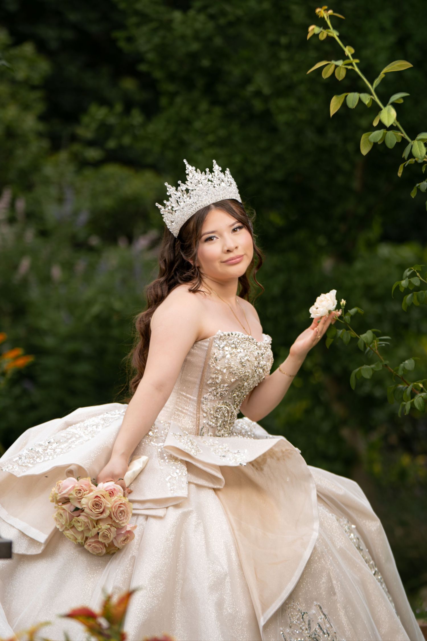 Young woman in elegant ballgown with crystal crown holds roses in a lush garden, resembling a fairytale princess.