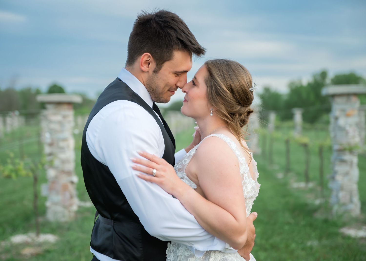 Bride and groom share an intimate moment, noses touching, embracing in a vineyard with stone pillars under a soft blue sky.