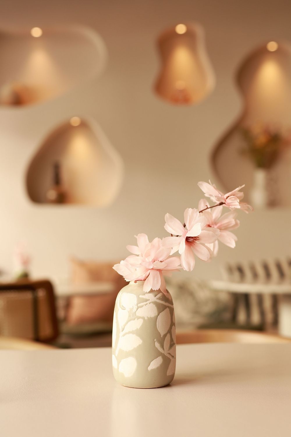 A ceramic vase with pink cherry blossoms on a white table, with warm glowing wall sconces in a soft beige interior.