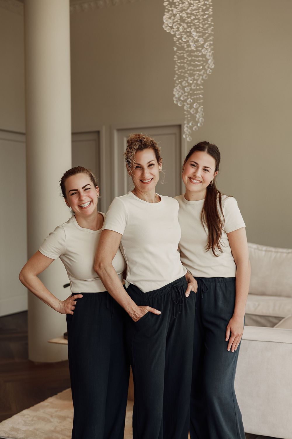 Three smiling women in matching white tops and dark pants pose together in an elegant modern interior.