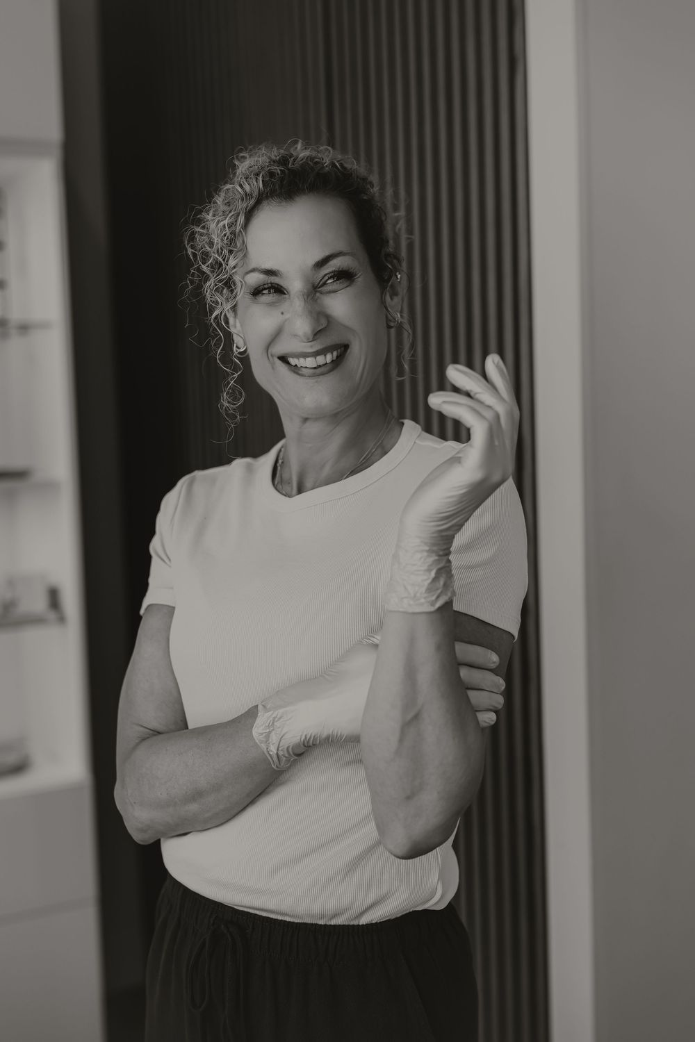 Black and white photo of a smiling woman in a white t-shirt putting on latex gloves in a clinical setting.