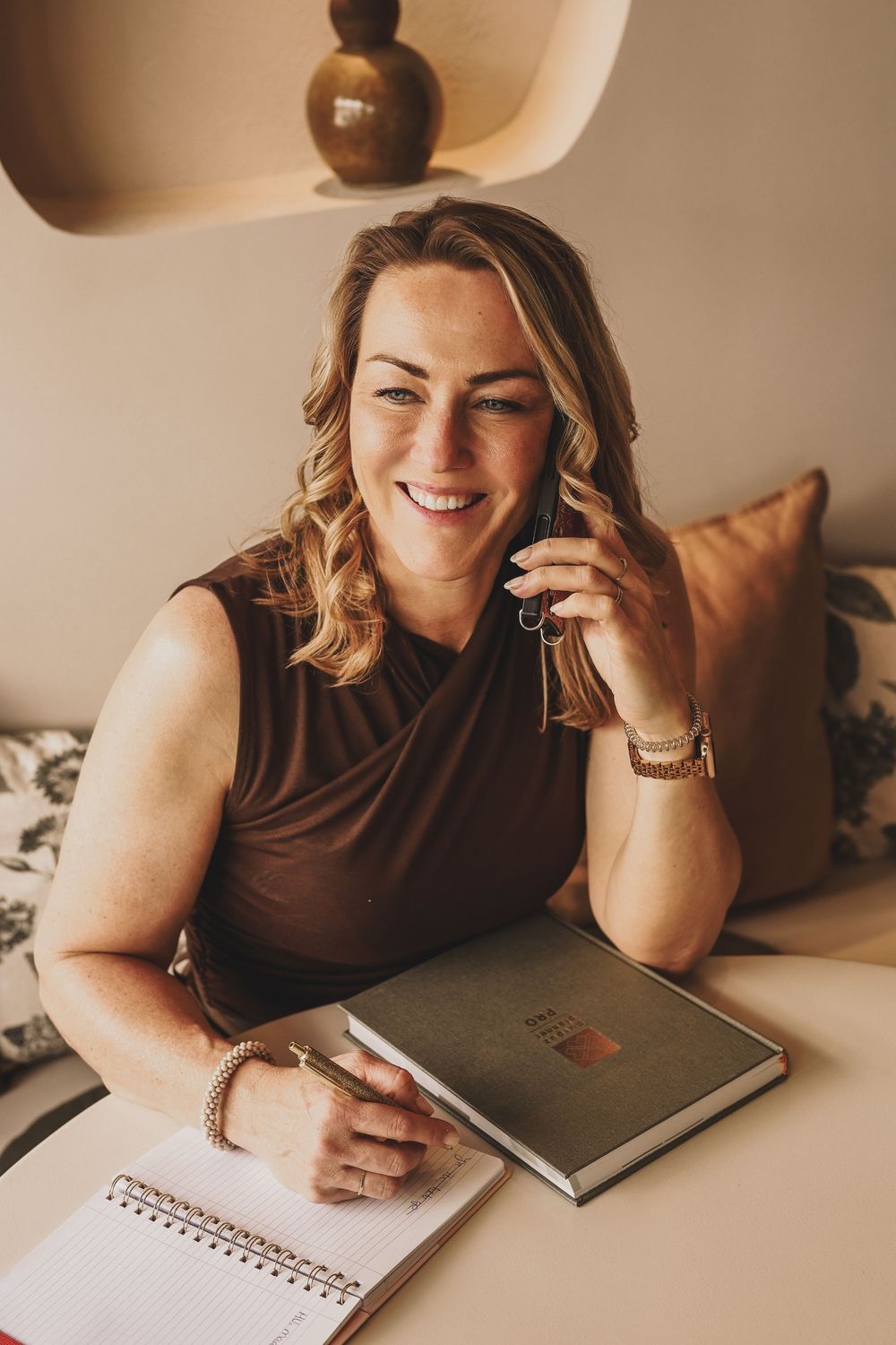 Smiling woman in brown dress sits at desk with notebook, talking on phone, wearing gold bracelets in warm neutral office.