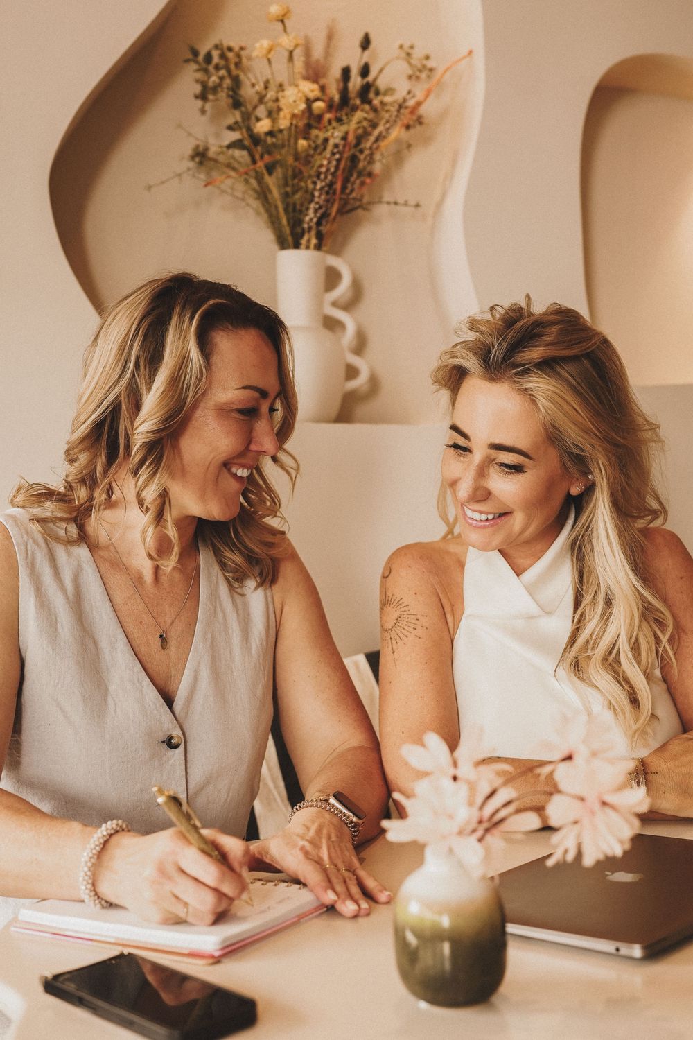 Two smiling blonde women collaborating at a table, writing in a notebook with a laptop and flowers nearby.