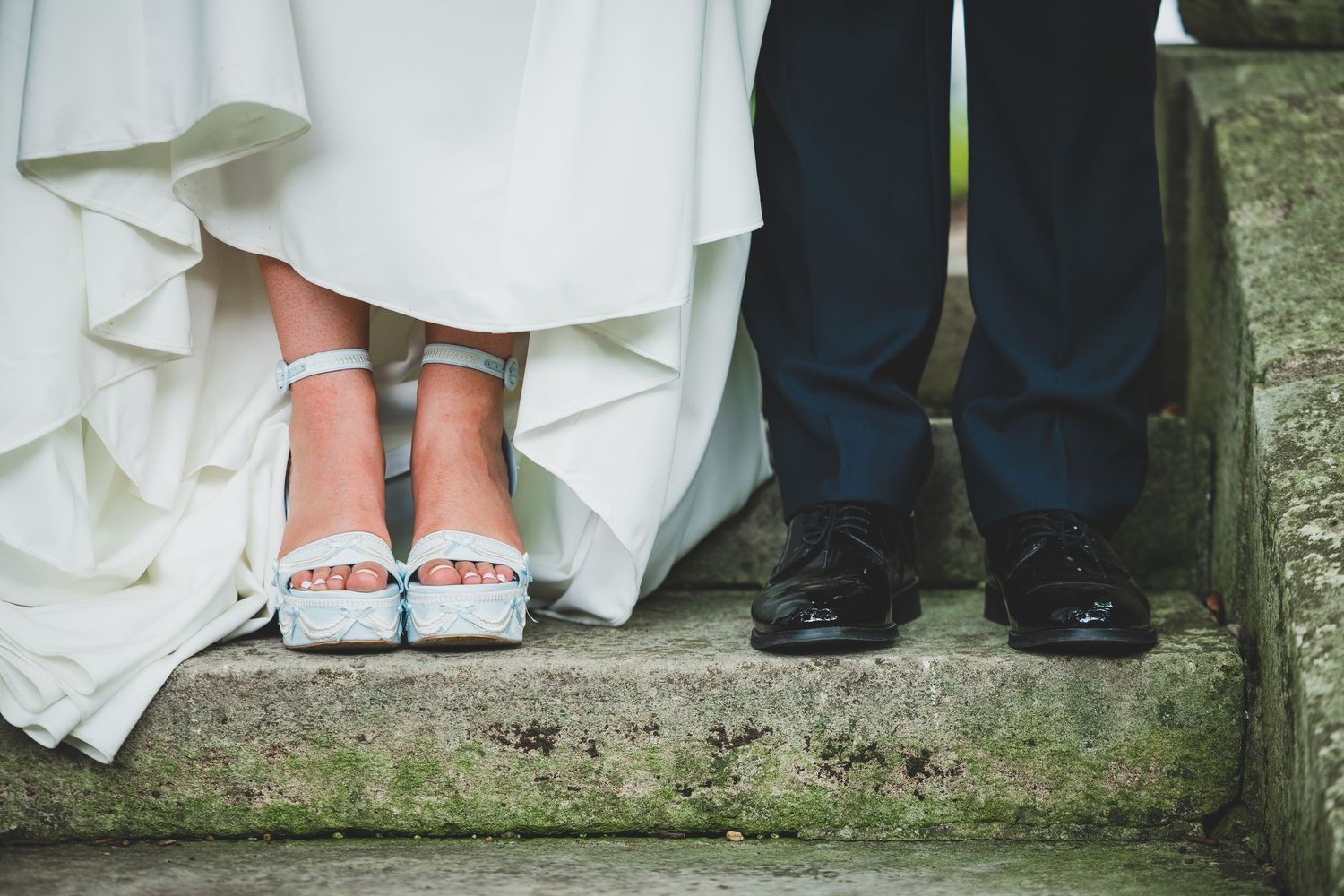 Close-up of bride in white dress wearing blue heels and groom in dark suit with black shoes standing on stone steps.