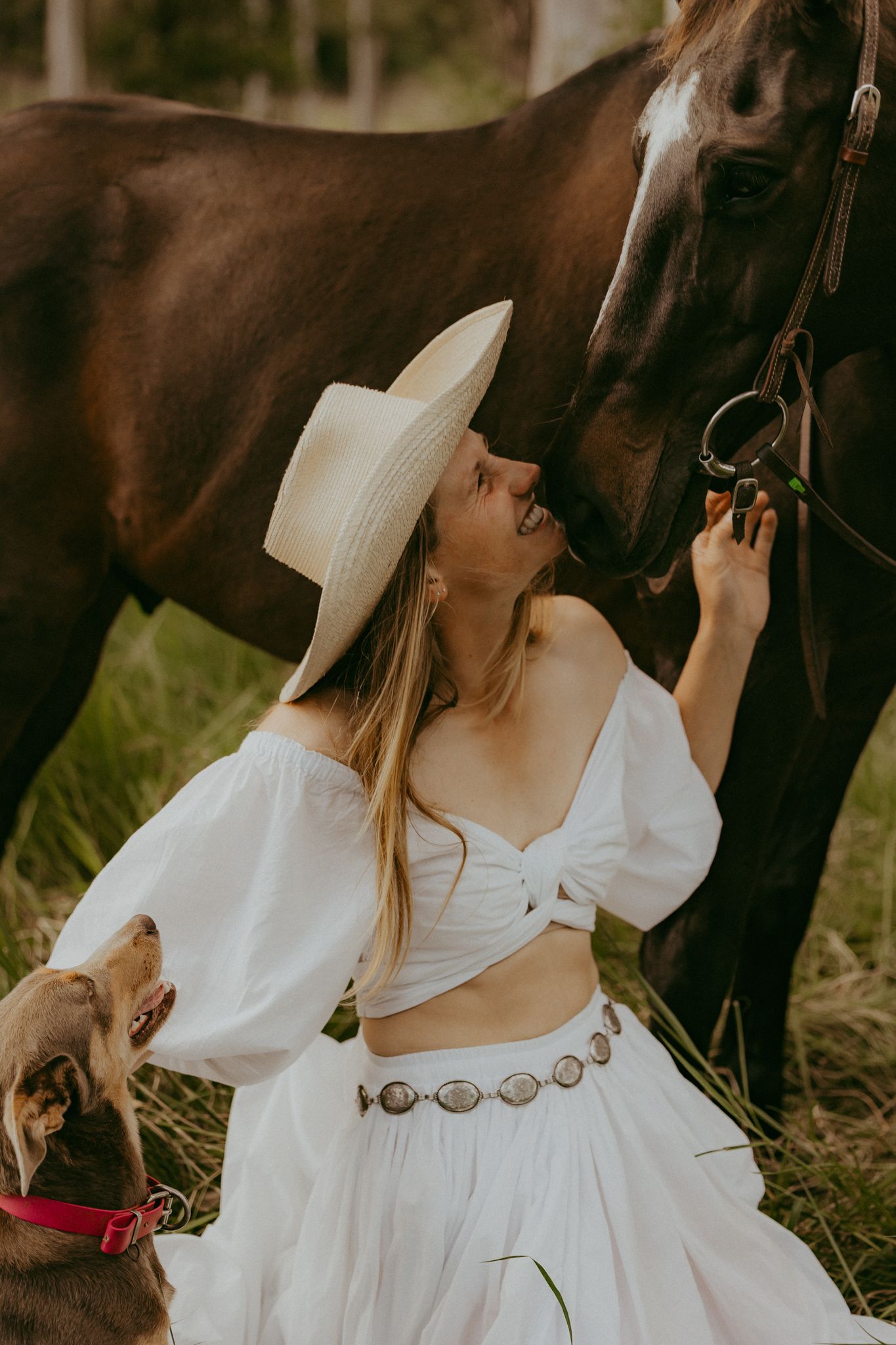 A blonde woman in a white off-shoulder outfit and hat sits beside a dark horse in a lush field.