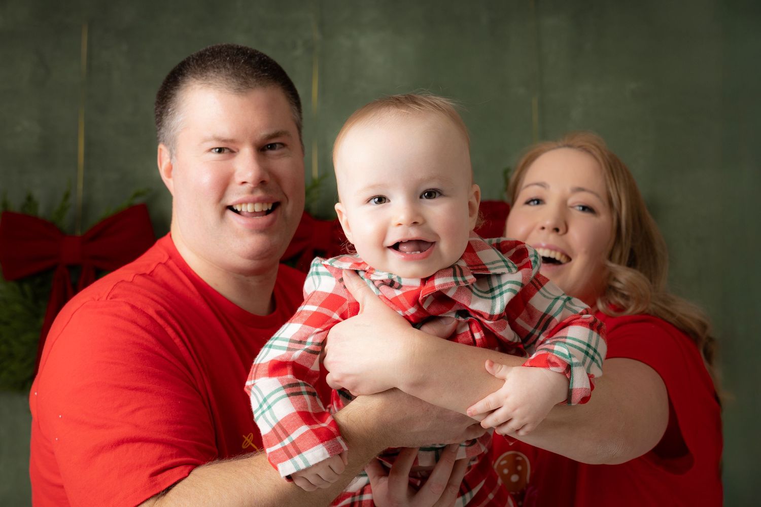 Happy family holiday portrait with parents in red shirts holding smiling baby in plaid outfit against green backdrop.