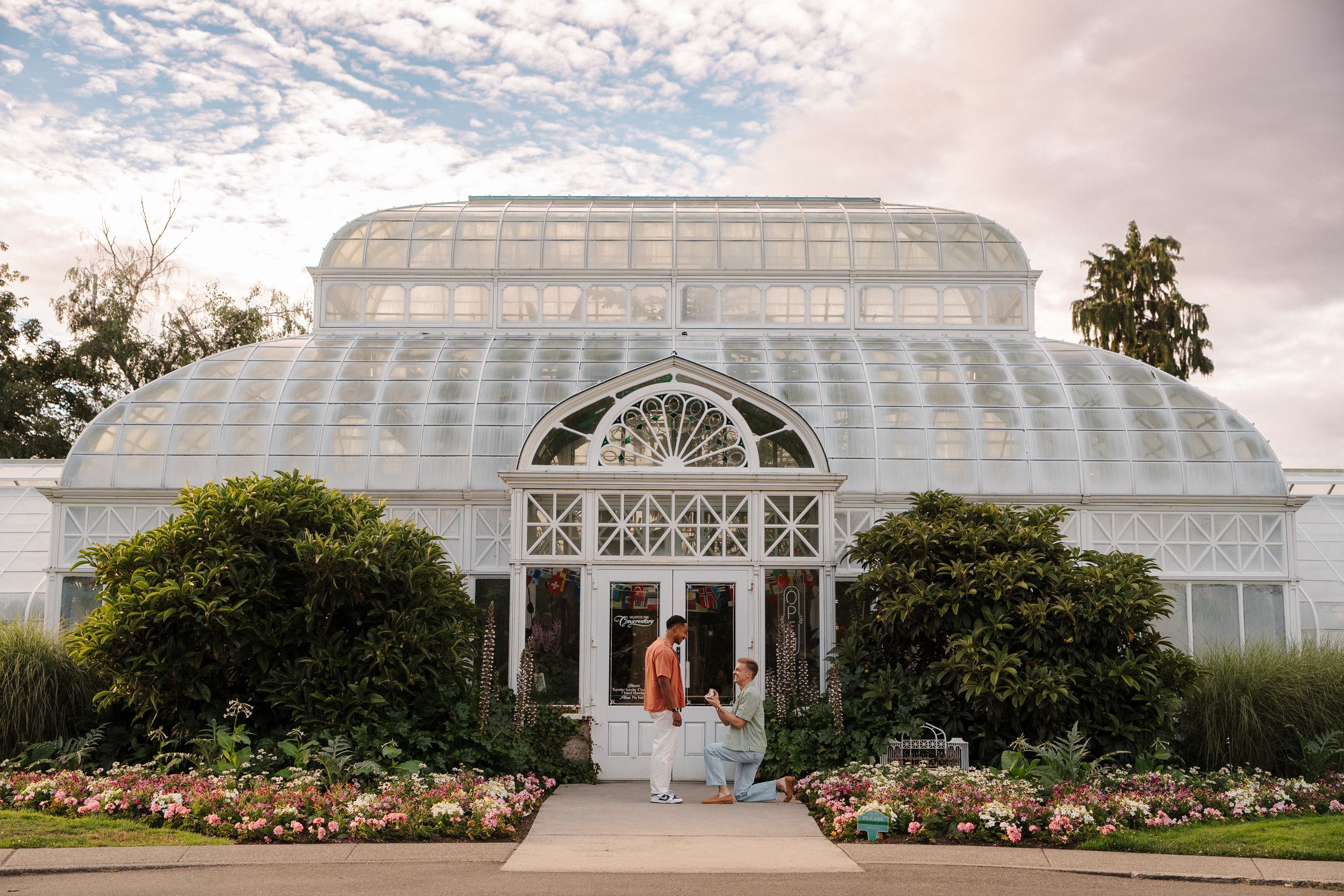 Couple poses in front of a grand Victorian glass greenhouse surrounded by colorful blooming gardens at dusk.