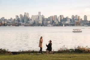 A man proposes to a woman by a waterfront park with a stunning city skyline backdrop at golden hour.