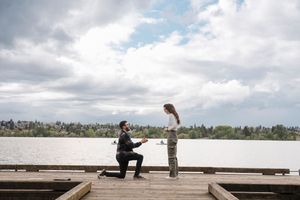 Man proposes on a wooden dock by a lake under cloudy skies, woman stands facing him in a romantic outdoor setting.