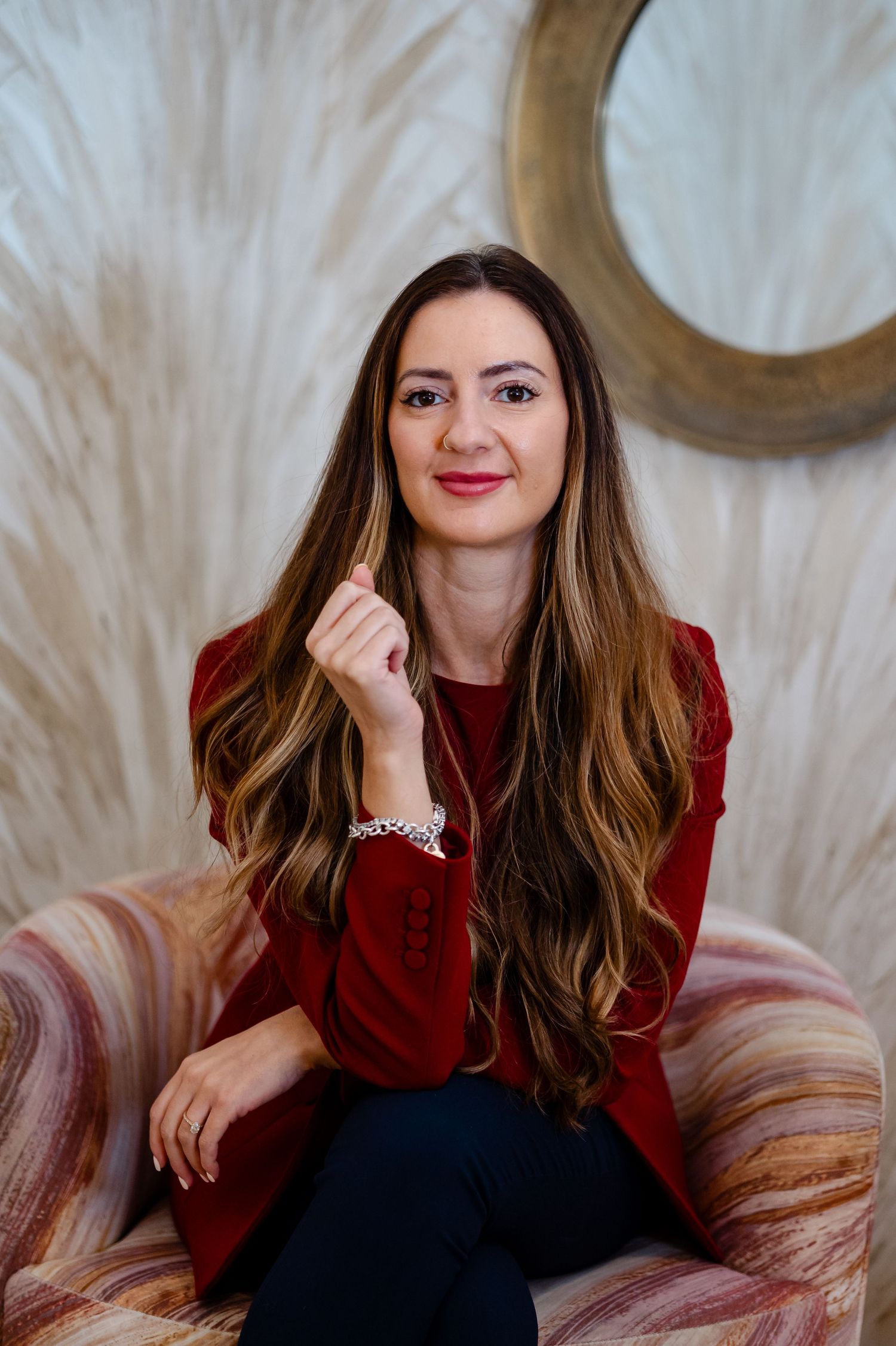 Woman with long brown hair in red blouse seated in colorful chair, posing confidently in elegant interior.