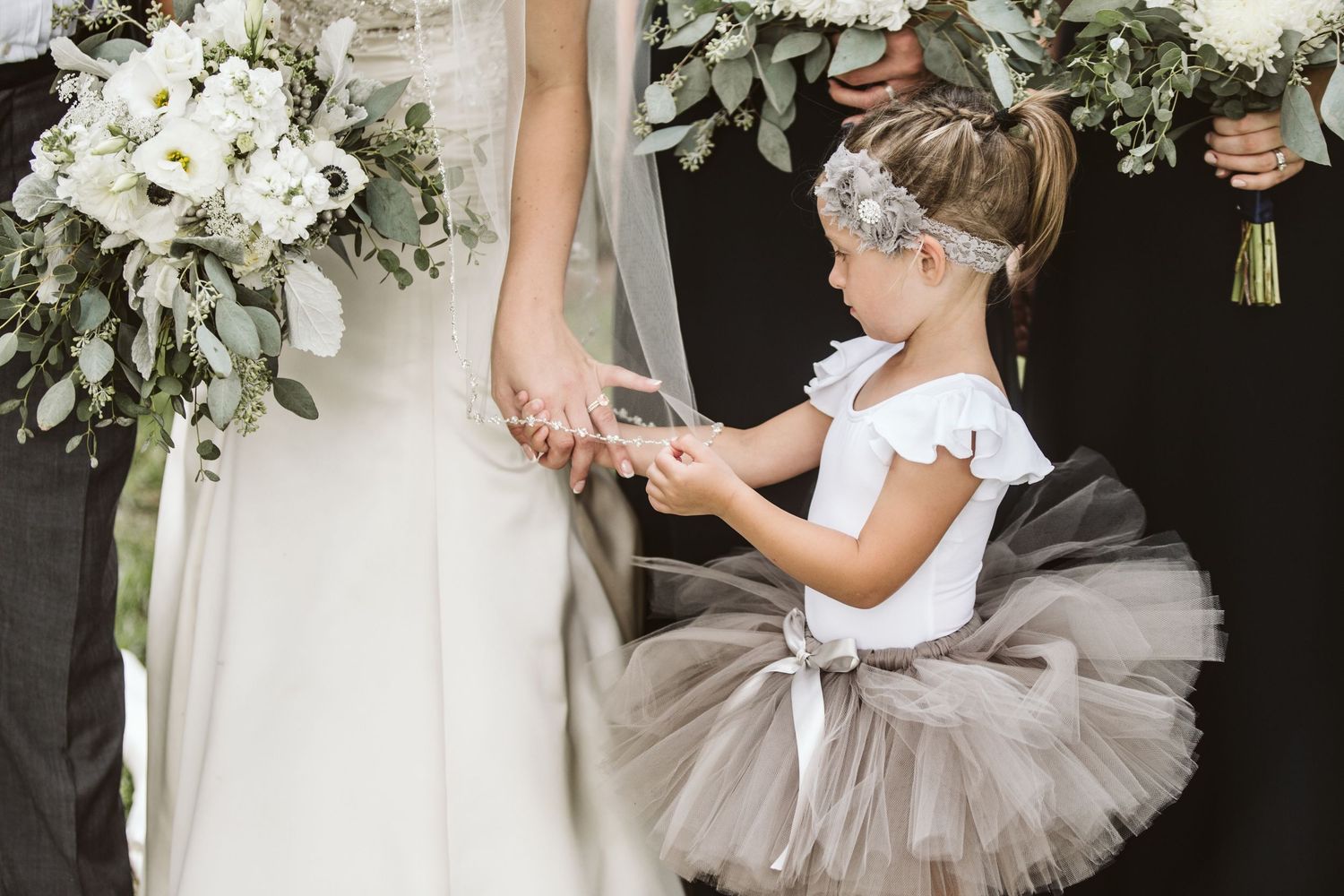 A flower girl in a gray tutu holds the bride's hand during a wedding ceremony with white floral bouquets.