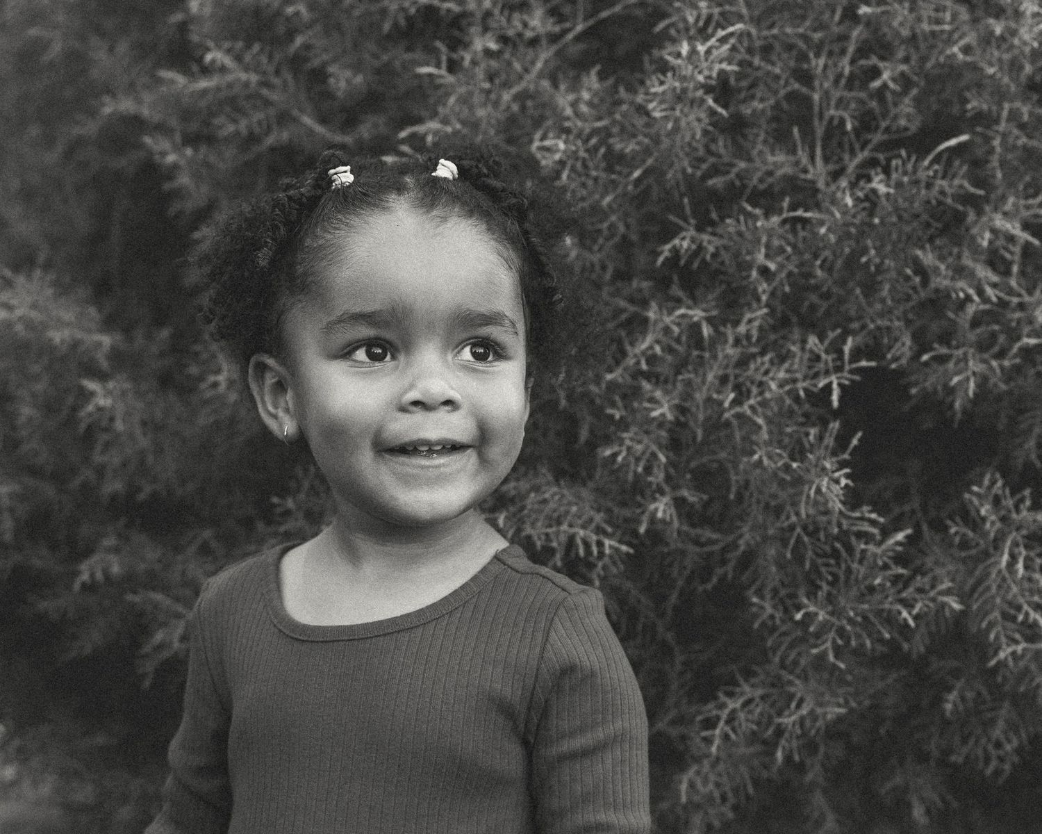 Black and white portrait of a smiling young girl with curly hair standing in front of lush green shrubs.