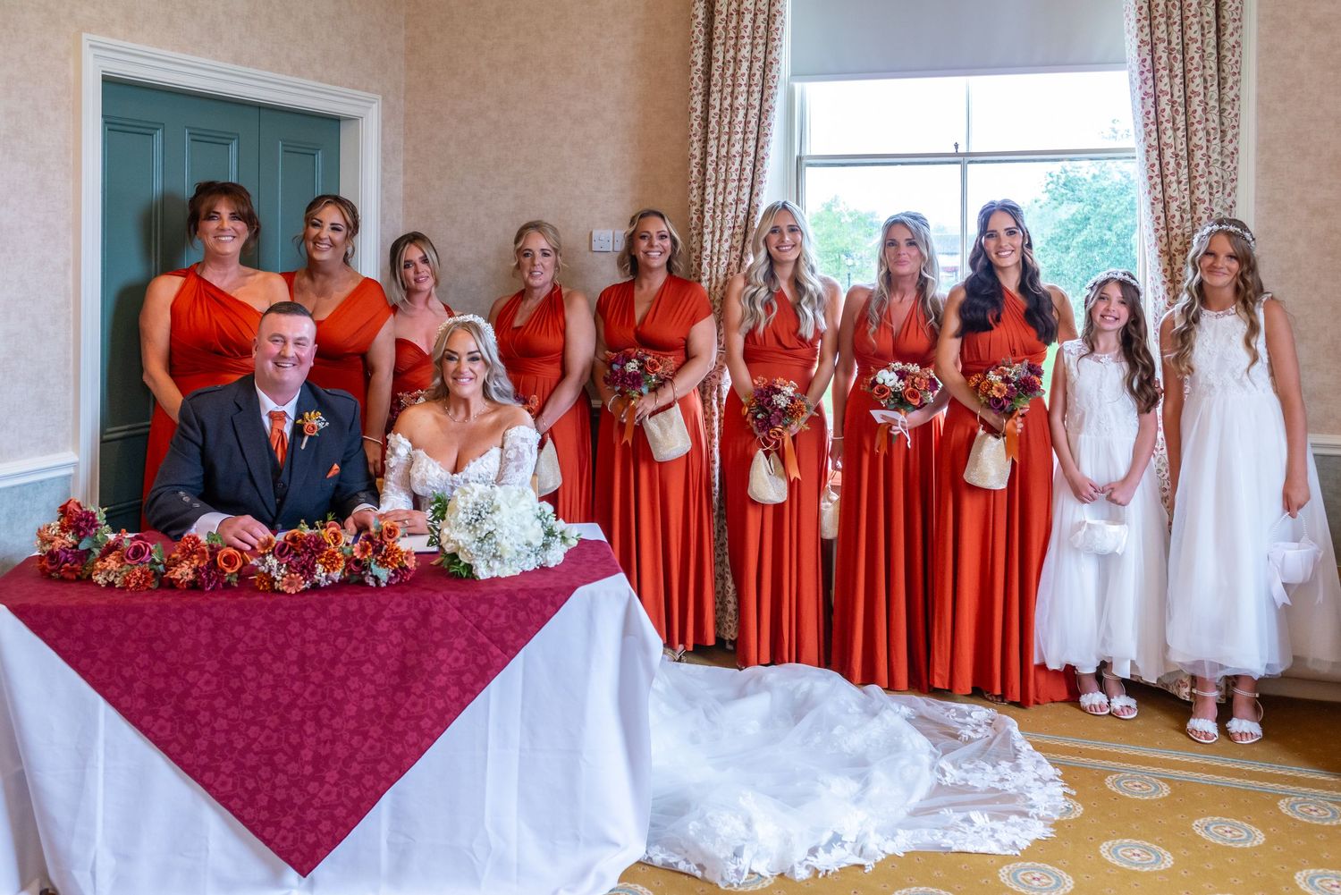 Bride and groom sign wedding register surrounded by bridesmaids in red dresses and flower girls in white at Runcorn Town Hall