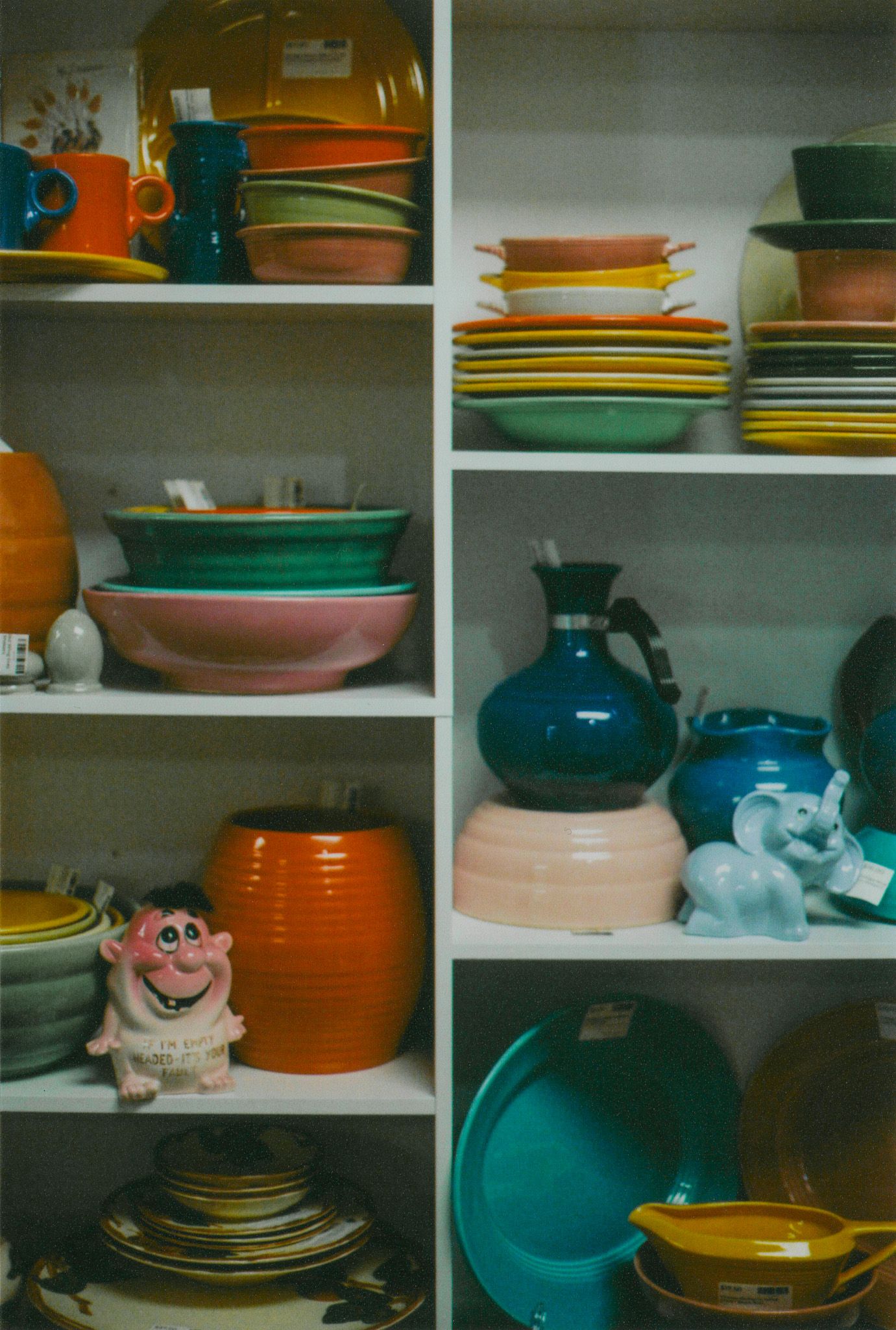 Vintage shelves filled with colorful Fiestaware dishes, bowls, pitchers, and an orange canister beside a pink ceramic pig and a blue elephan