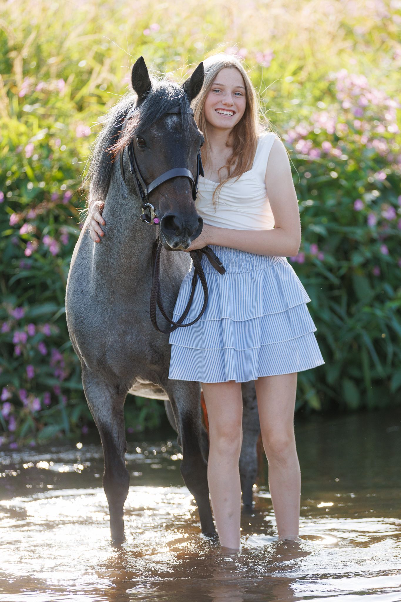Woman in white lace dress walks alongside a brown horse on a sunlit forest path.