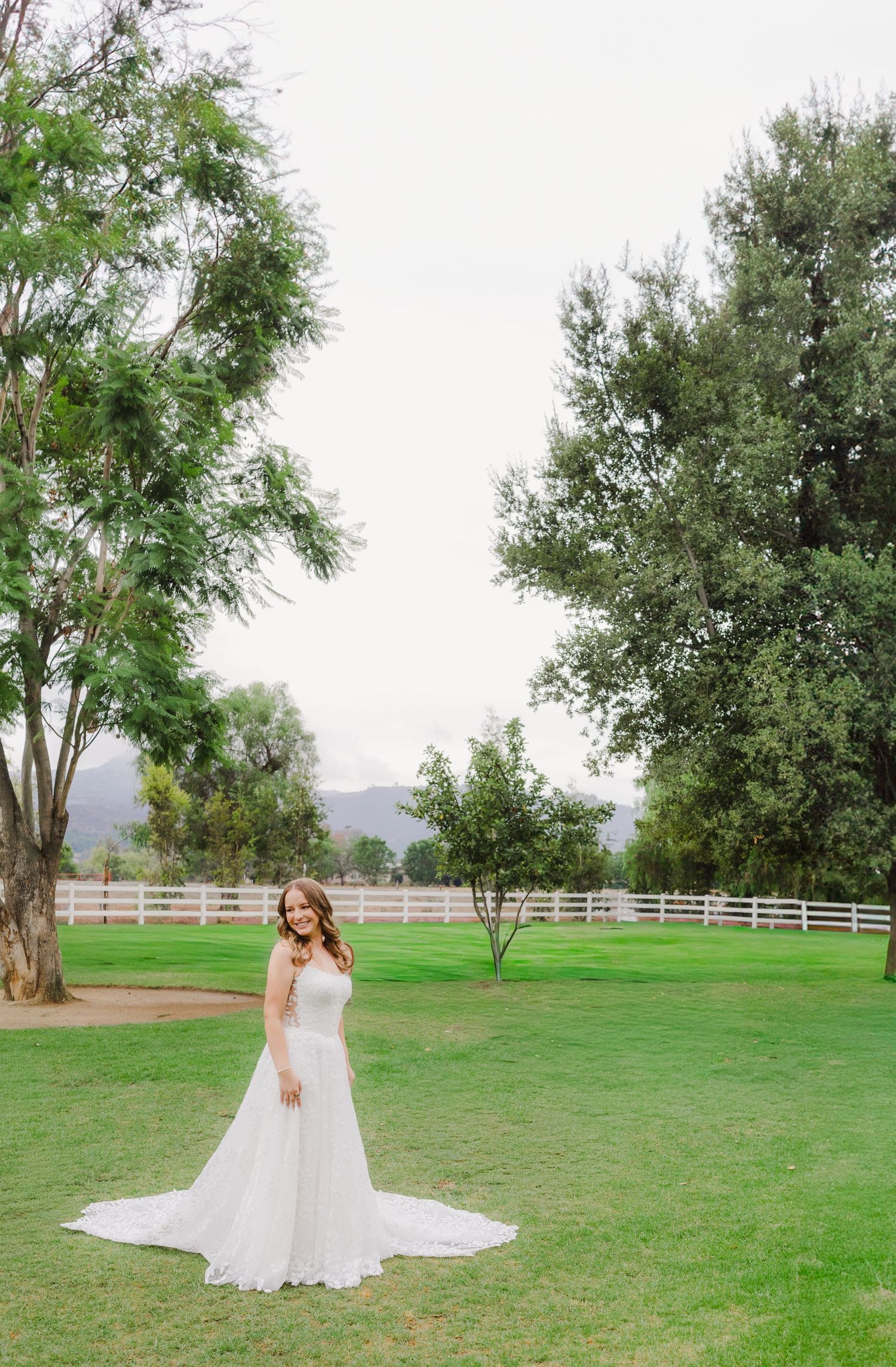 Bride in a flowing lace wedding gown stands on a green lawn framed by tall trees, a white fence and distant mountains.