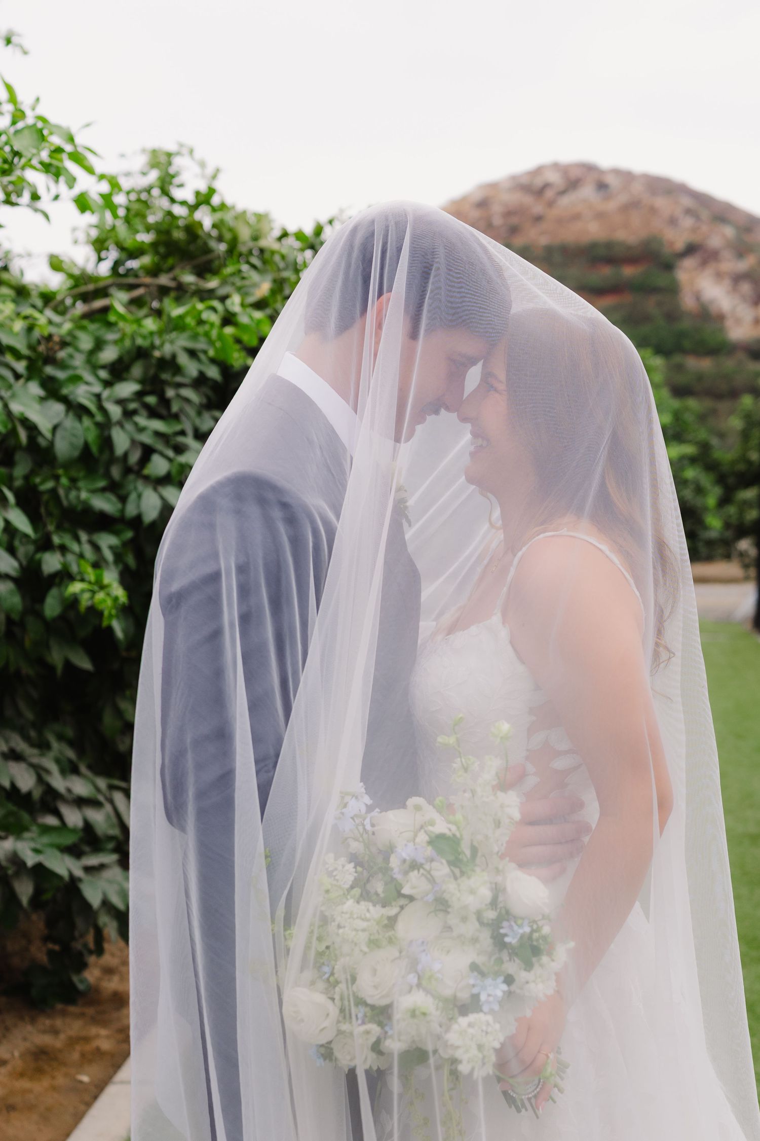 Bride and groom touch foreheads and smile under a sheer wedding veil in a garden, with a bridal bouquet held in front of them.