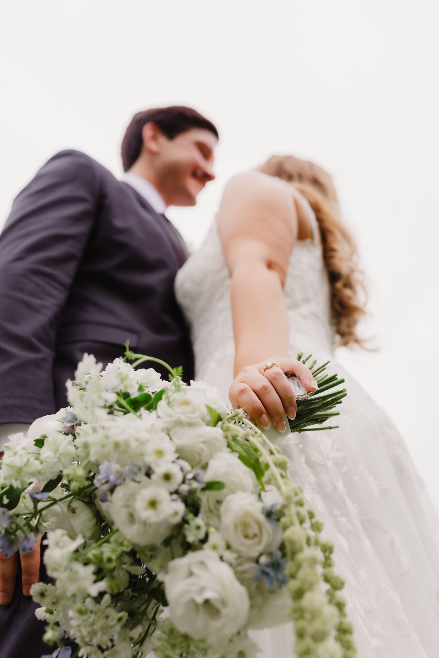 Low-angle view of a smiling bride and groom with a white and blue floral bouquet in the foreground and the bride's ring visible.