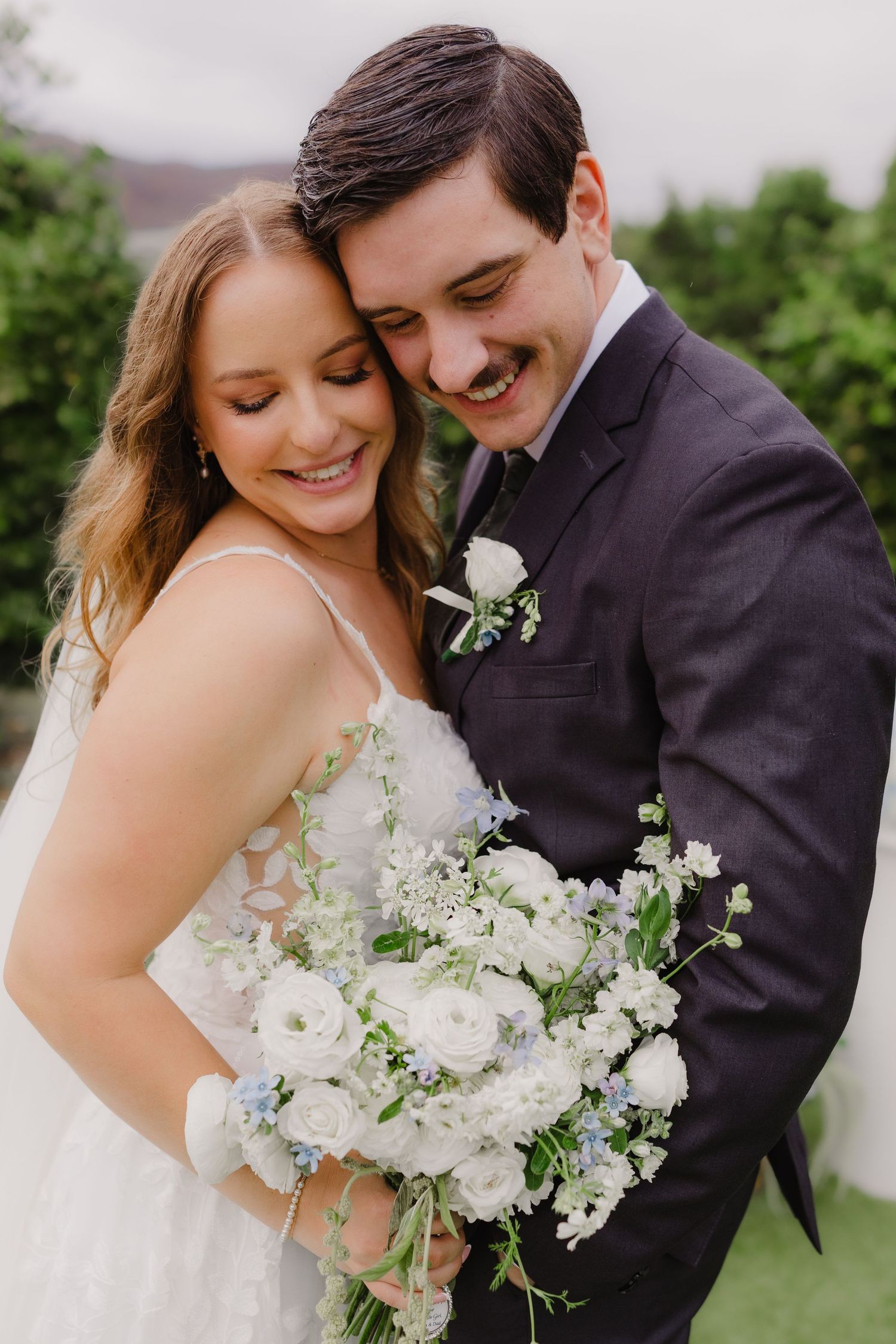 Happy bride and groom embrace closely outdoors, smiling while holding a white and blue wedding bouquet of roses and wildflowers.
