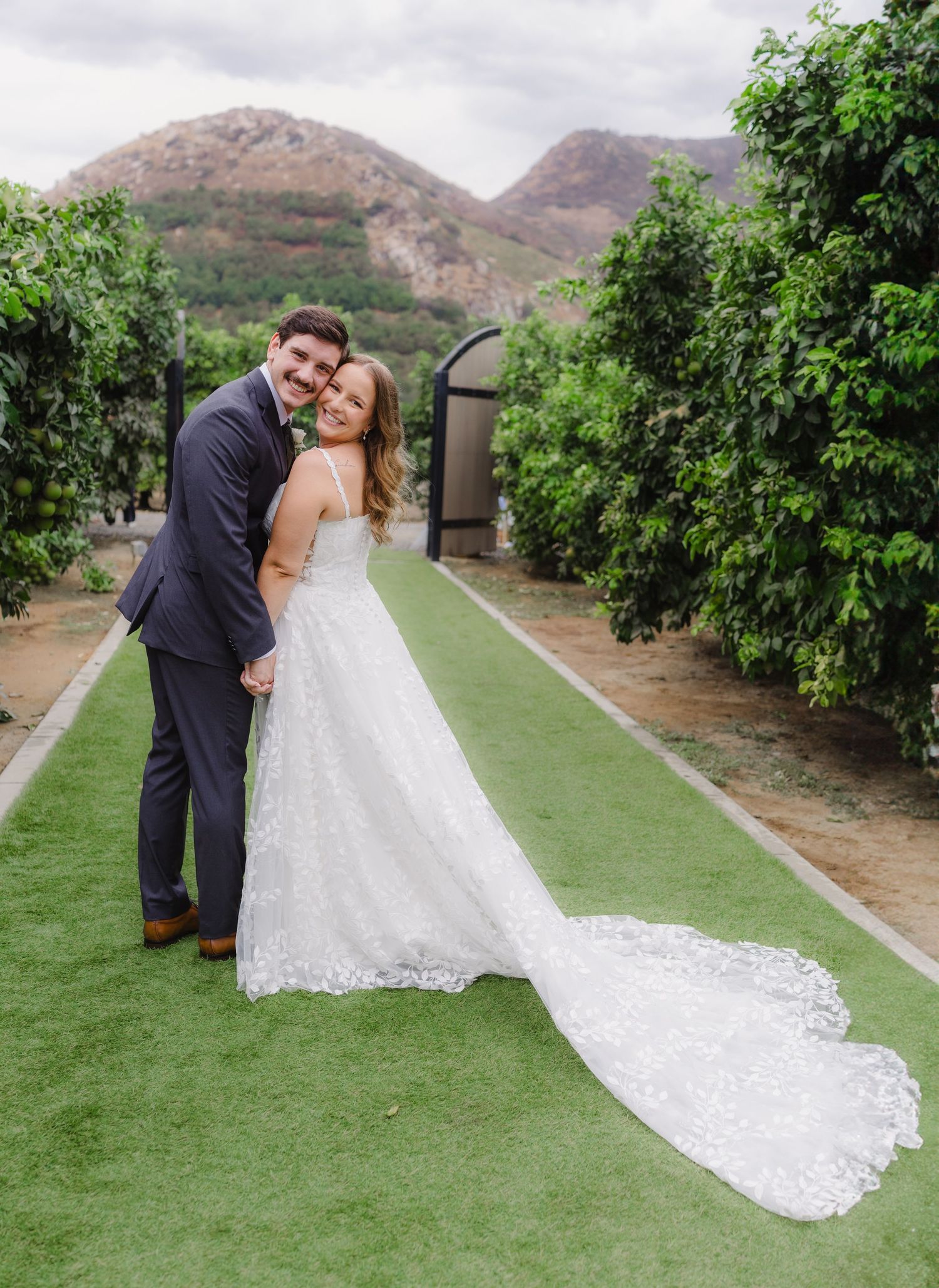 Smiling bride and groom pose on a green lawn aisle with a long lace train, citrus trees and mountains in the background.
