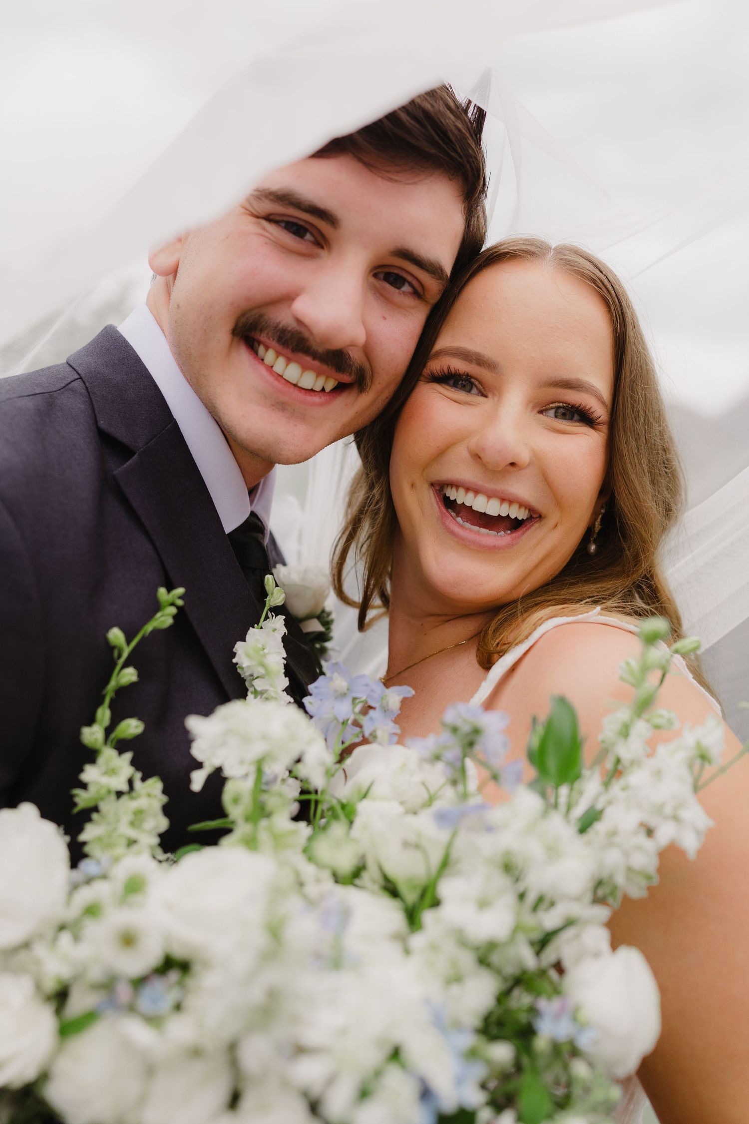 Close-up of a joyful bride and groom laughing under a veil, holding a bouquet of white and blue wedding flowers.